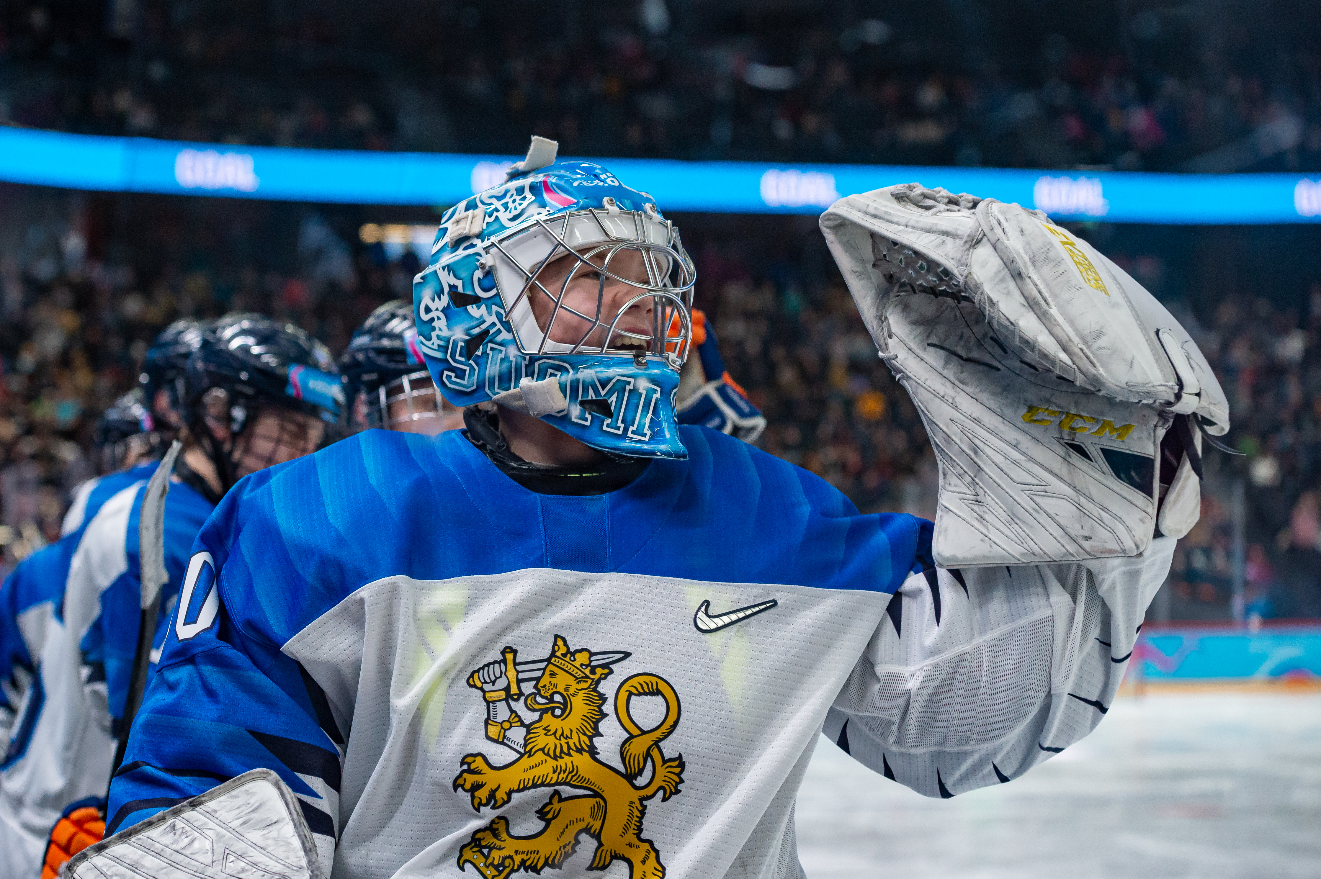 Niklas Kokko celebrates a teammate’s goal with glove hand up in the air, donning the uniform of Team Finland and a blue goalie mask that says “Suomi” along the bottom