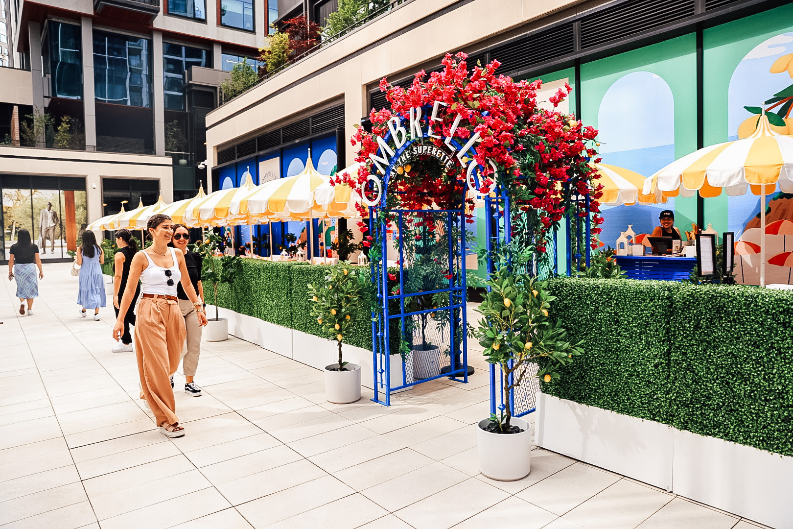 A blue trellis covered with pink flowers holds signage that says Ombrello. It leads into a colorful sidewalk patio setup on a city street.