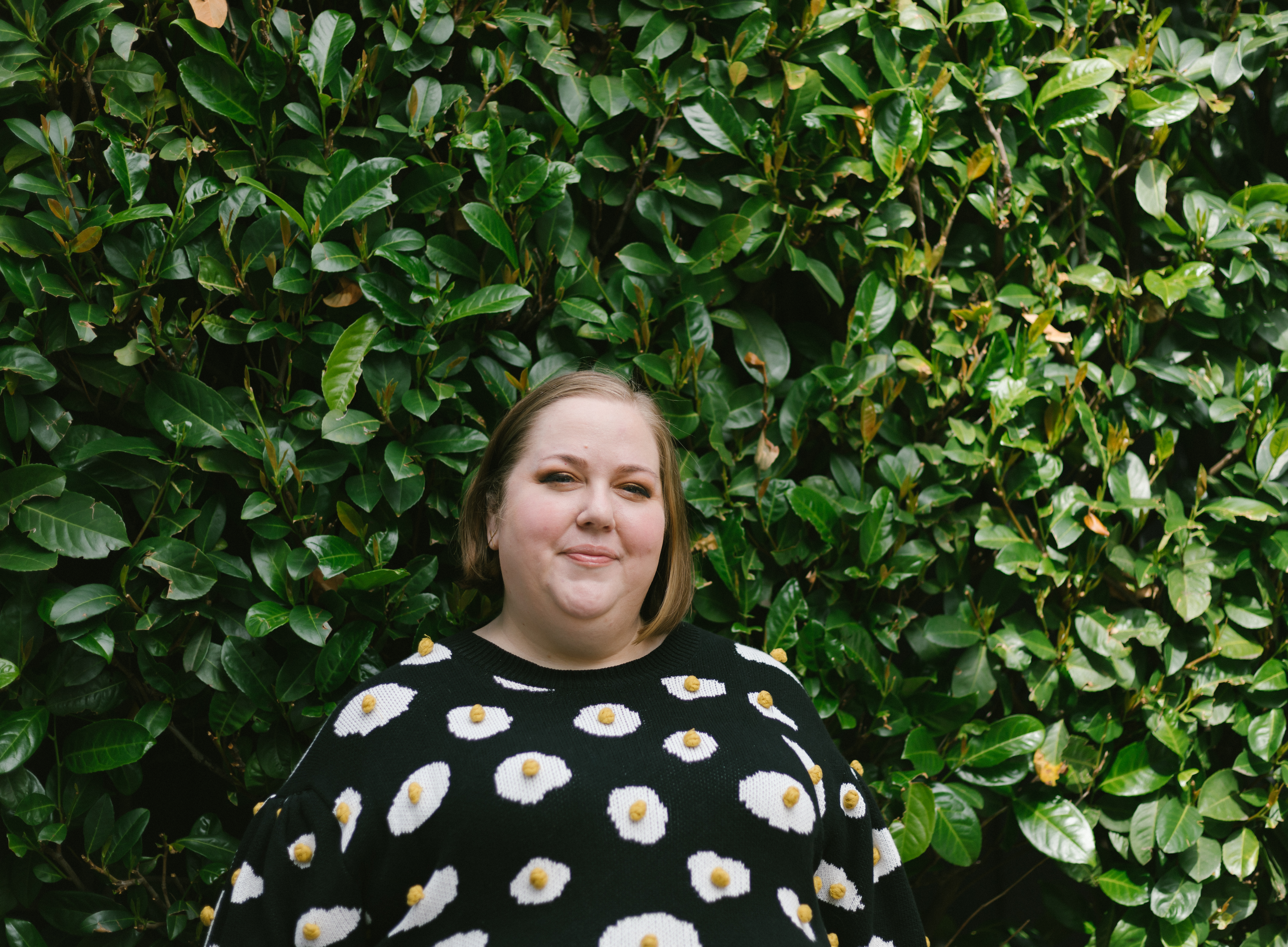 Podcaster and writer Aubrey Gordon stands in an egg sweater in front of a green hedge.