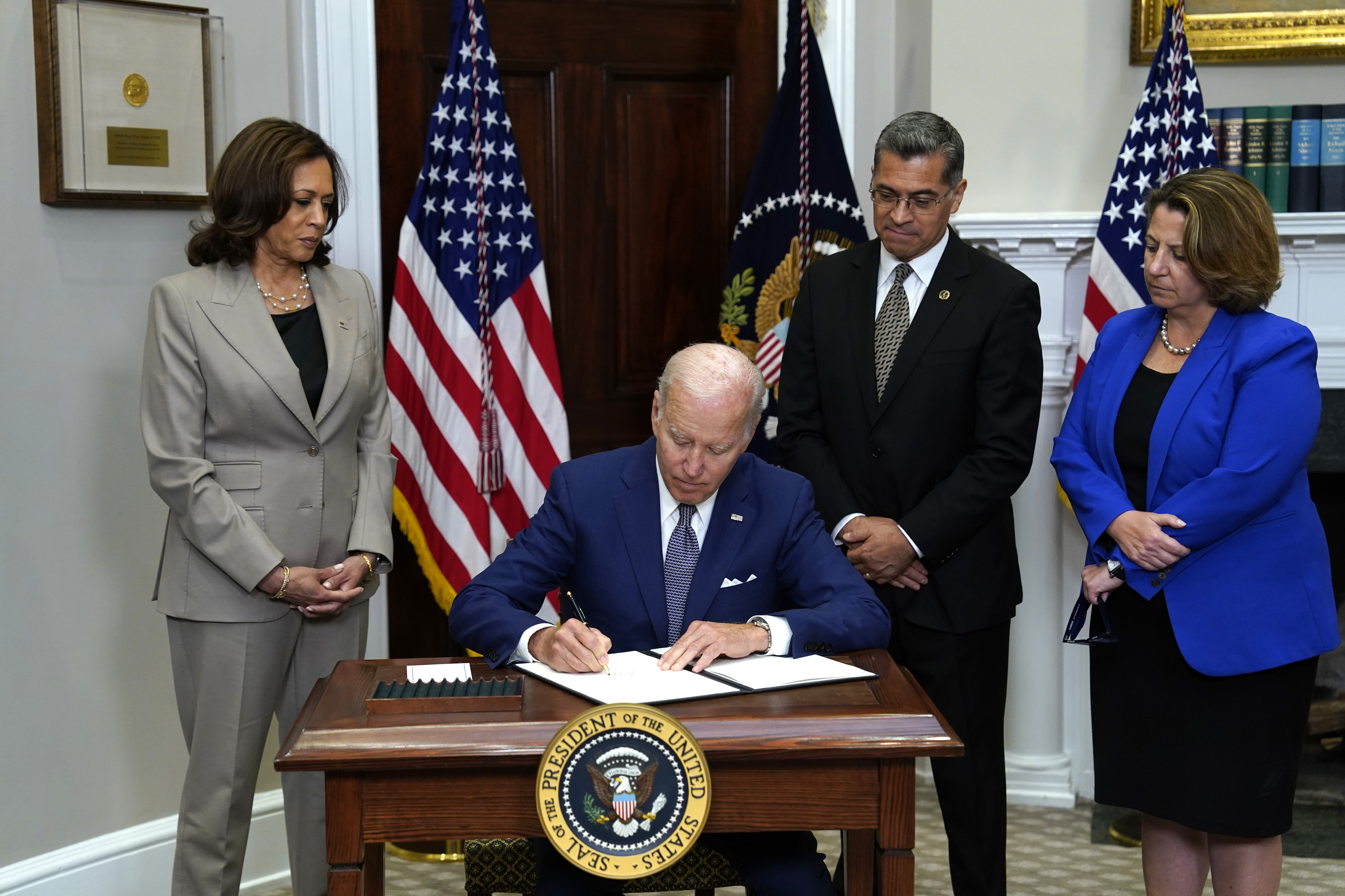 President Biden sits and signs at a desk, while Vice President Kamala Harris, 