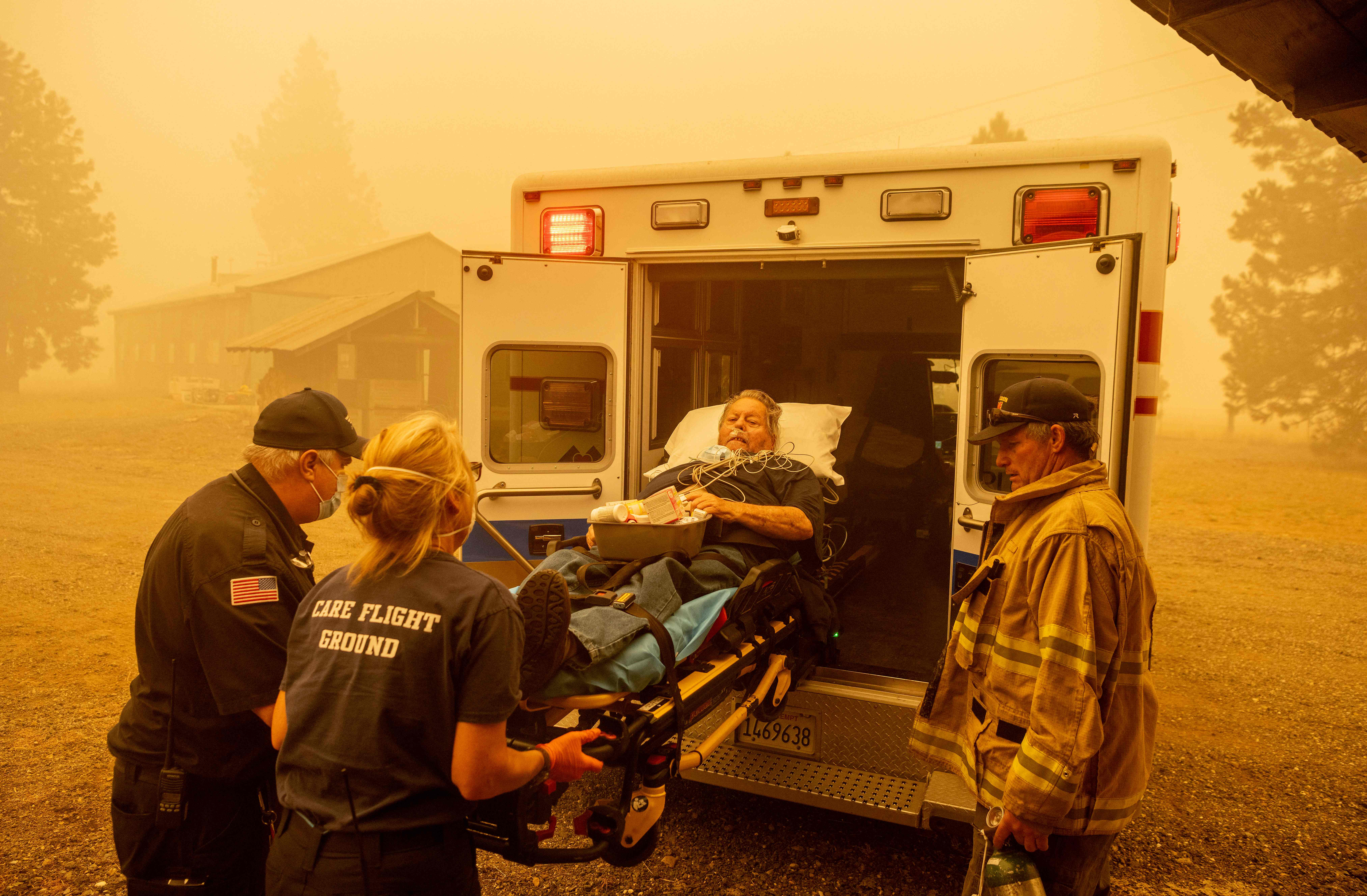 A man on a gurney is rushed into an ambulance by EMTs, with voluminous smoke and orange-tinted air from wildfires surrounding them.