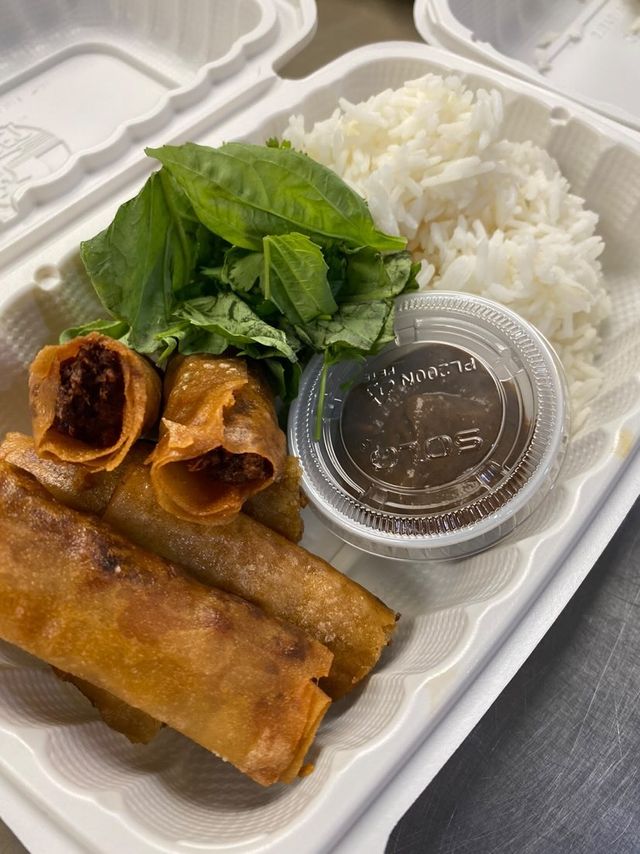A styrofoam container with fried egg rolls and greens and ice and a container of dark sauce.