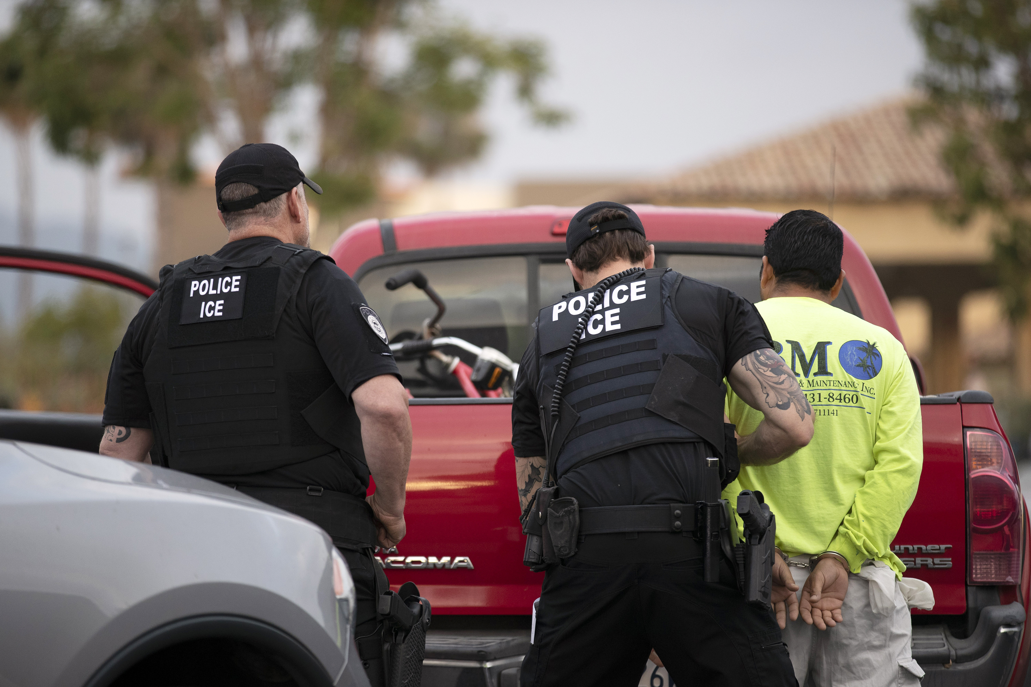 Two immigration officers escort a person in handcuffs past a red pickup truck.
