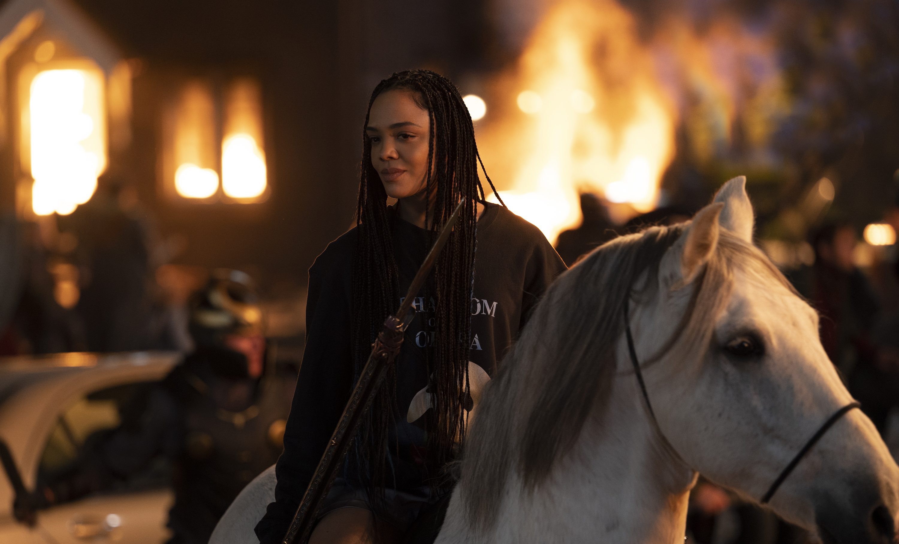 Tessa Thompson as Valkyrie in Thor: Love and Thunder, sitting on horseback in a night scene lit by burning buildings in the background