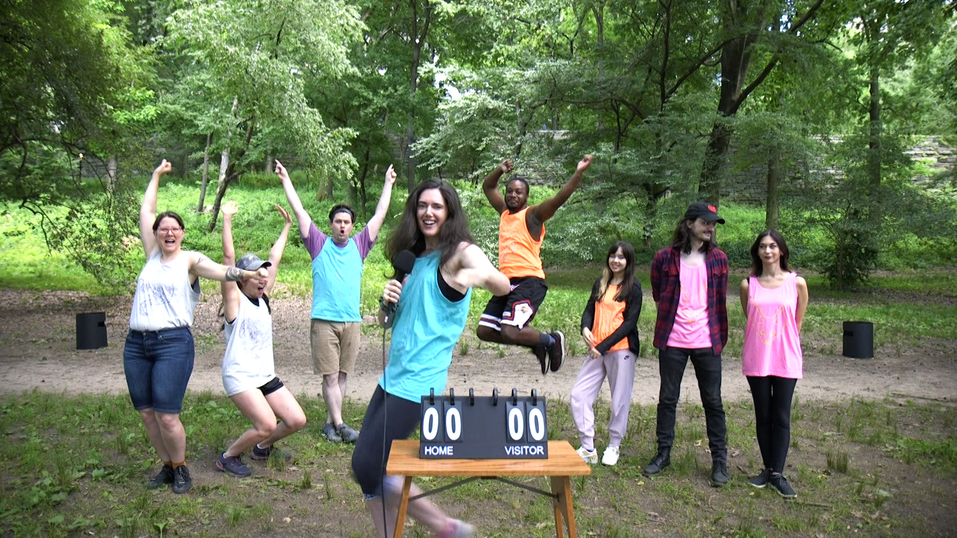 Eight people stand in a park with KanJam cans behind them and a scoreboard in front of them. Some of the people are cheering or jumping in the air.