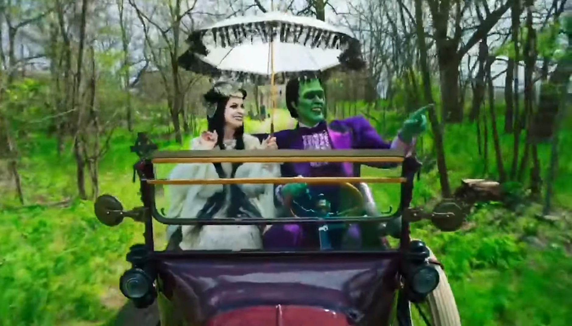 The Munsters - Frankenstein’s Monster and Lily, a vampire, ride in a fancy purple car through a bright green wood. Lily is dressed in white with an elaborate headpiece and parasol, and Herman Munster is pointing at something off screen. They are having a pleasant time