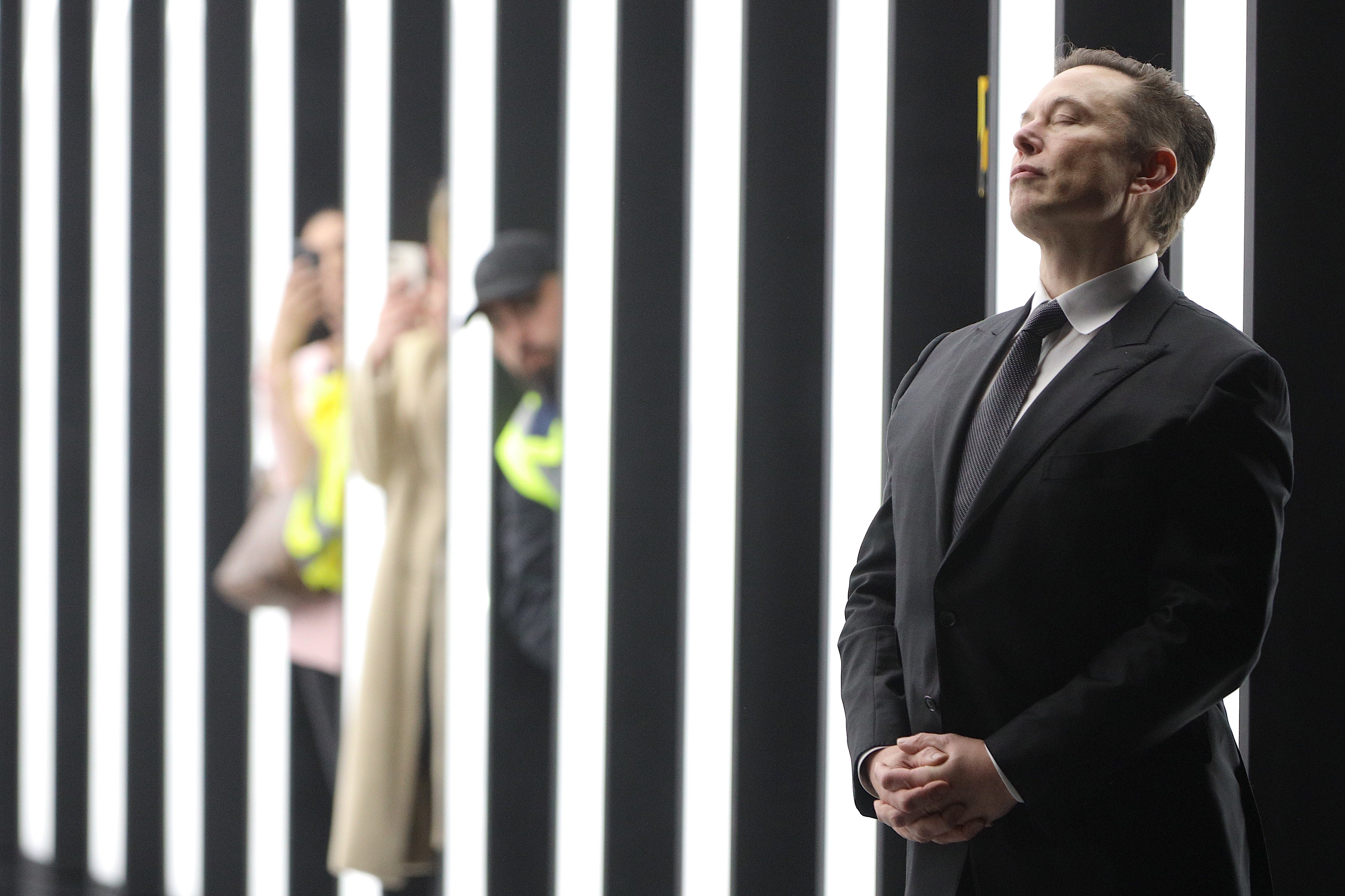 Elon Musk stands in front of tunnel of bright light at the new Tesla manufacturing plant in Germany, while workers peek at him from behind.