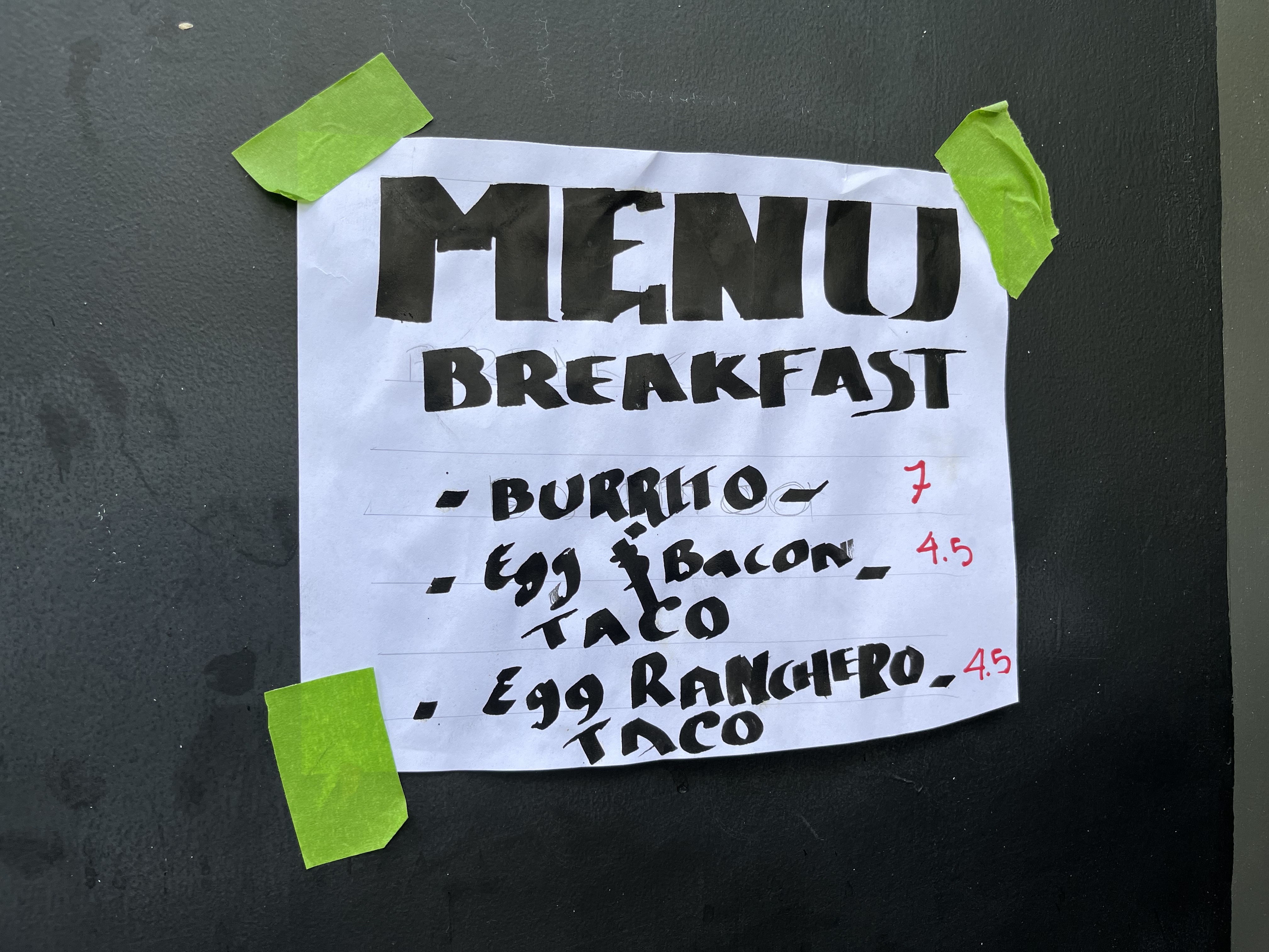 A white sheet of paper taped to a wall with green tape. It has the word MENU written in large capitals, followed by Breakfast, followed by Burrito; Egg bacon taco; and egg ranchero taco.