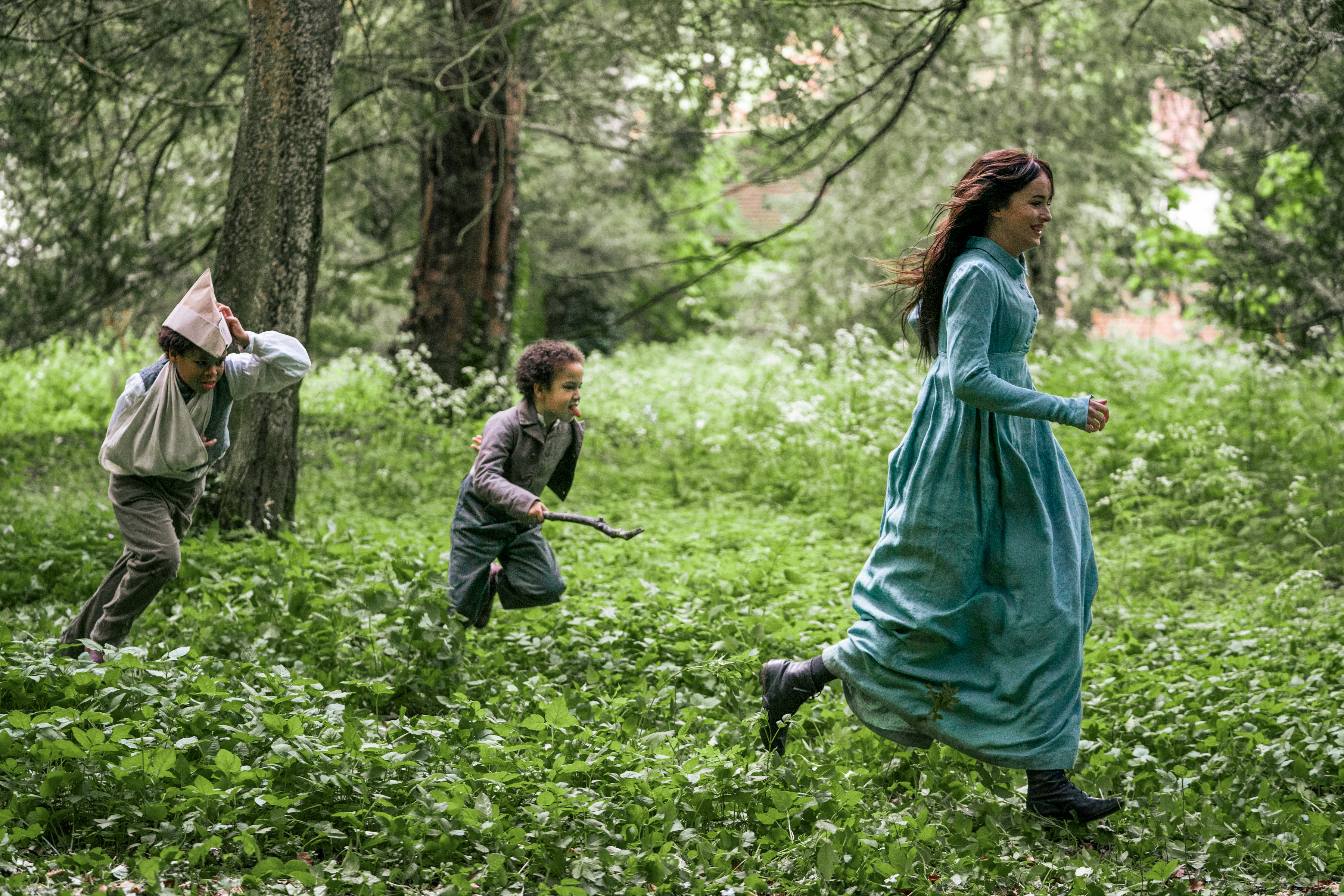 A group of children in paper hats chase a woman in a long Regency gown through the forest. They are all laughing.