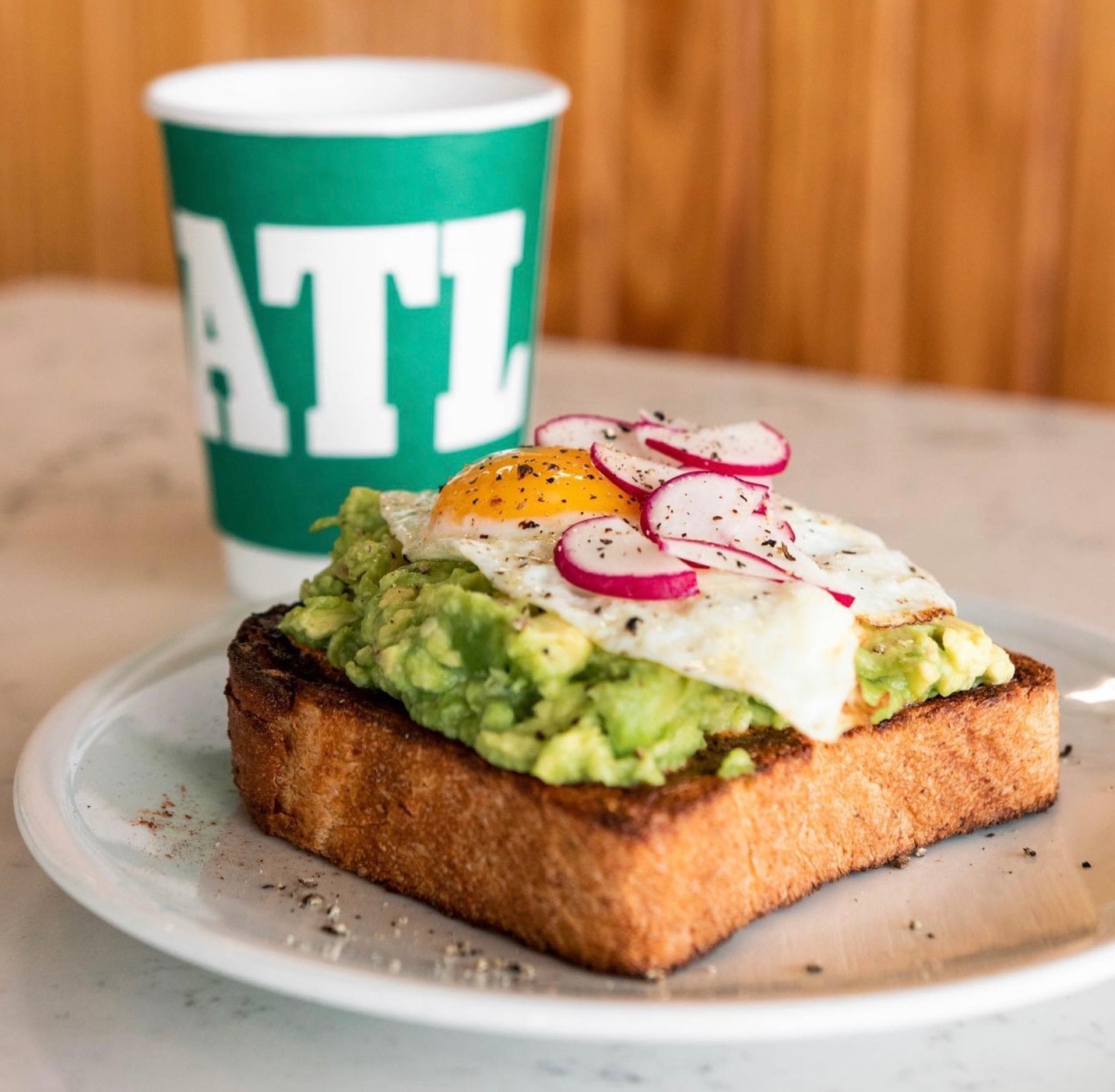 A thick slice of toast topped with avocado spread, a sunny side egg and slice radishes with a green to-go cup of coffee with the letters ATL from Spiller Park Coffee in Atlanta.