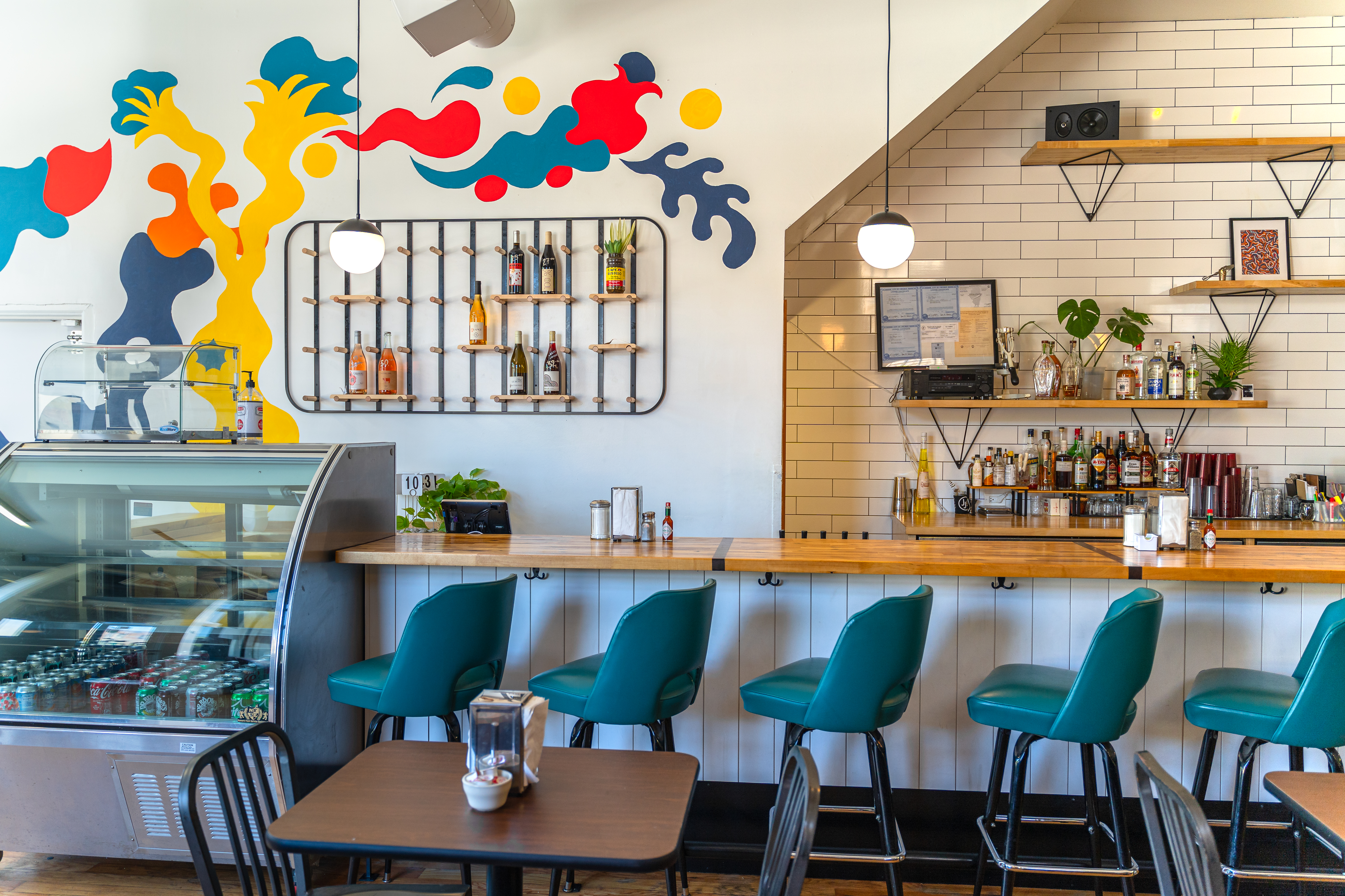 A white restaurant space with a diner counter lined by teal stools. A colorful, abstract mural is painted on one wall.
