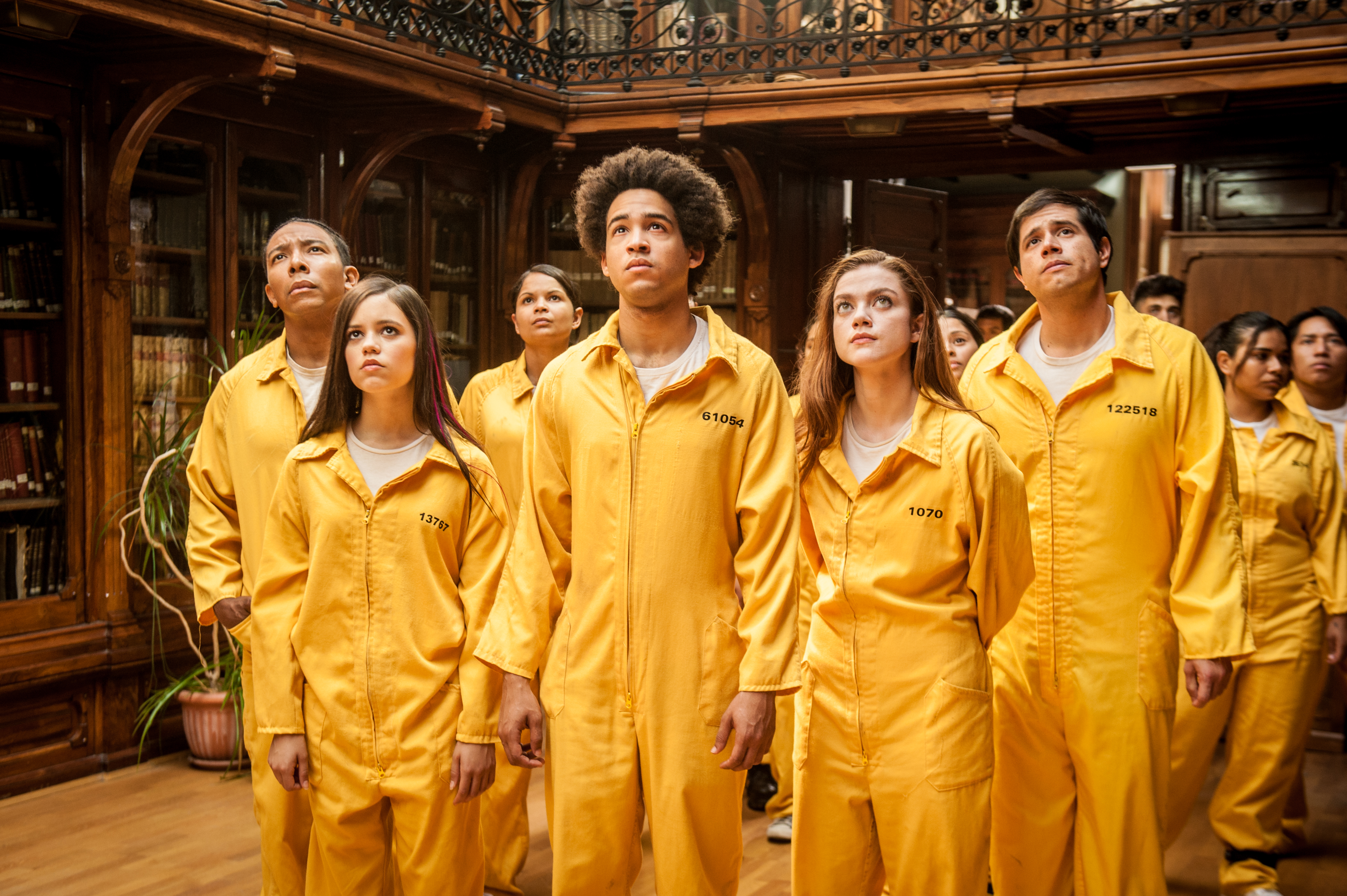 The cast of American Carnage stands in a library wearing yellow jumpsuits looking at someone above them