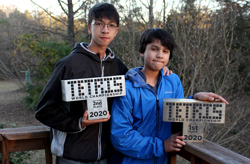 A 15-year-old boy standing next to a 13-year-old boy. Each of them is holding a statue for 2020’s Classic Tetris World Championship.