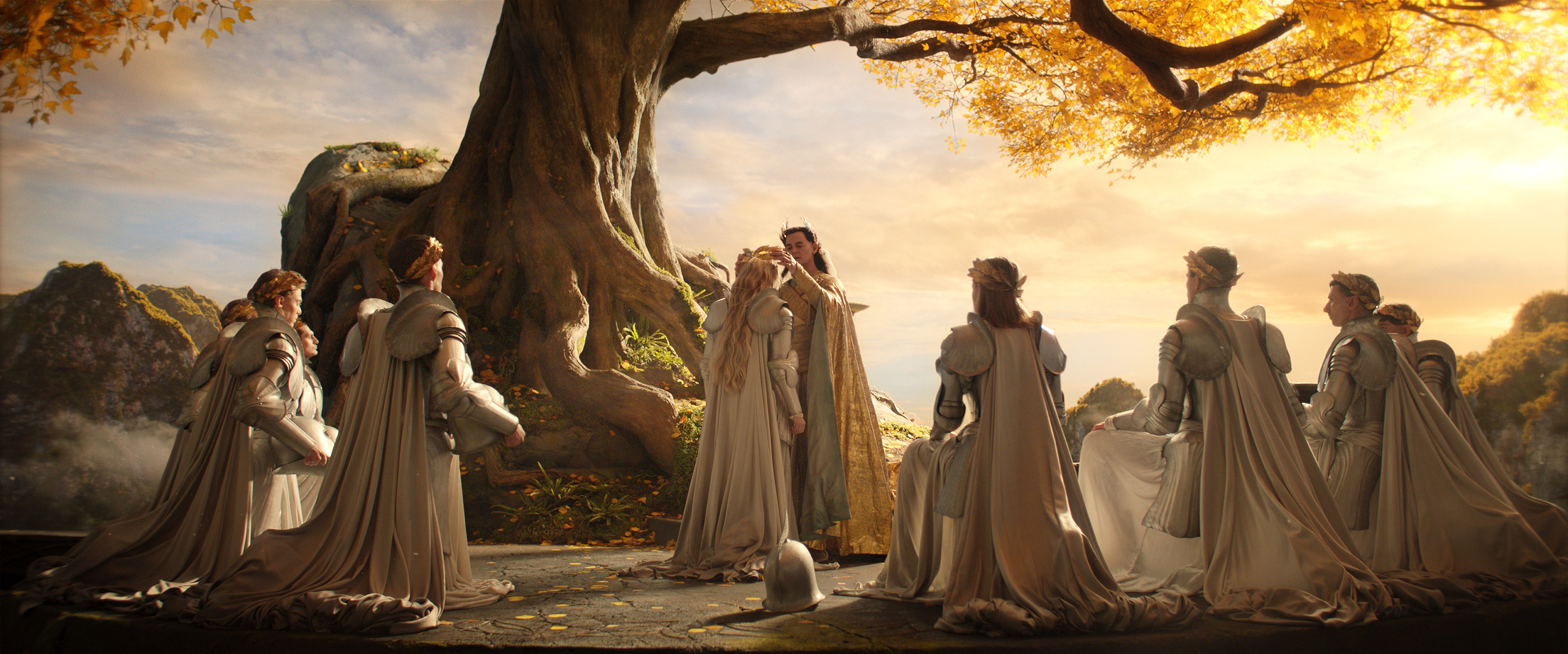Gil-galad bestowing a crown upon Galadriel’s head as eight other elves look on, sitting in a circle around them, next to a big tree, in The Lord of the Rings: The Rings of Power