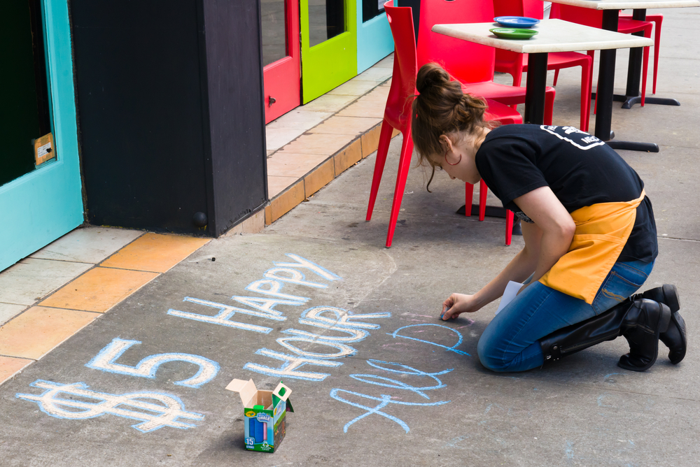 A server writes in chalk on the sidewalk outside of a bar advertising $5 happy hour drink specials.