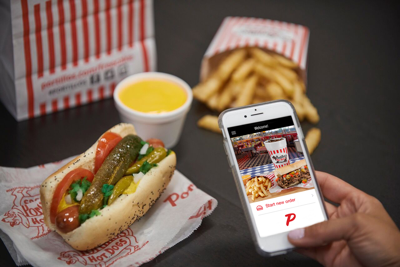 A Chicago-style hot dog from Portillo’s lies in relief, next to an order of fries and dipping sauce. In the foreground, a hand holding a phone places an order.
