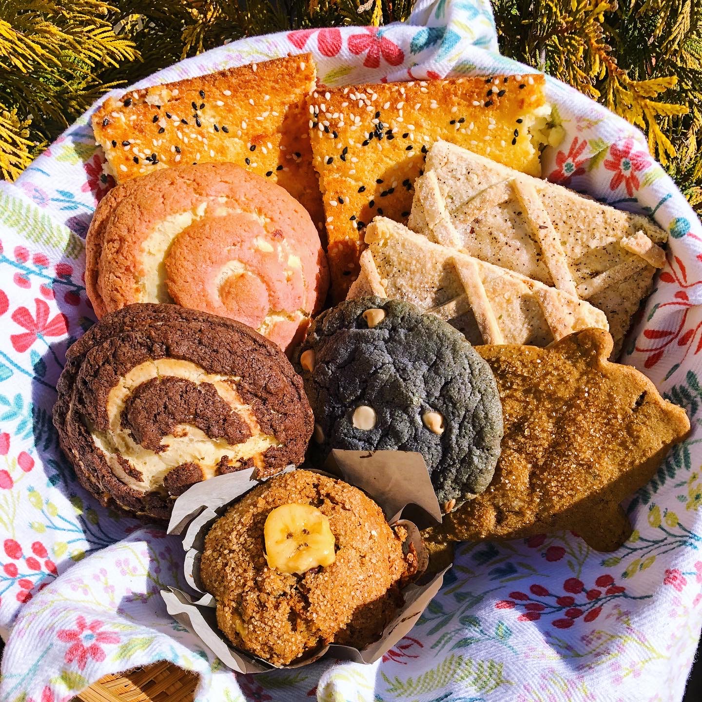 A basket of pan dulce nestled in the grass. A white tea towel beneath the pastries with a pink flower pattern. In the back, two golden square pastries with black and white sesame seeds scattered across the surface. Two other rectangular-shaped pastry has a criss-cross texture. A chocolate cookie in the center, surrounded by round, swirled pan dulce dusted with cinnamon. In the foreground is a pan dulce with a single banana slice baked into the center. Warm light illuminates the basket.