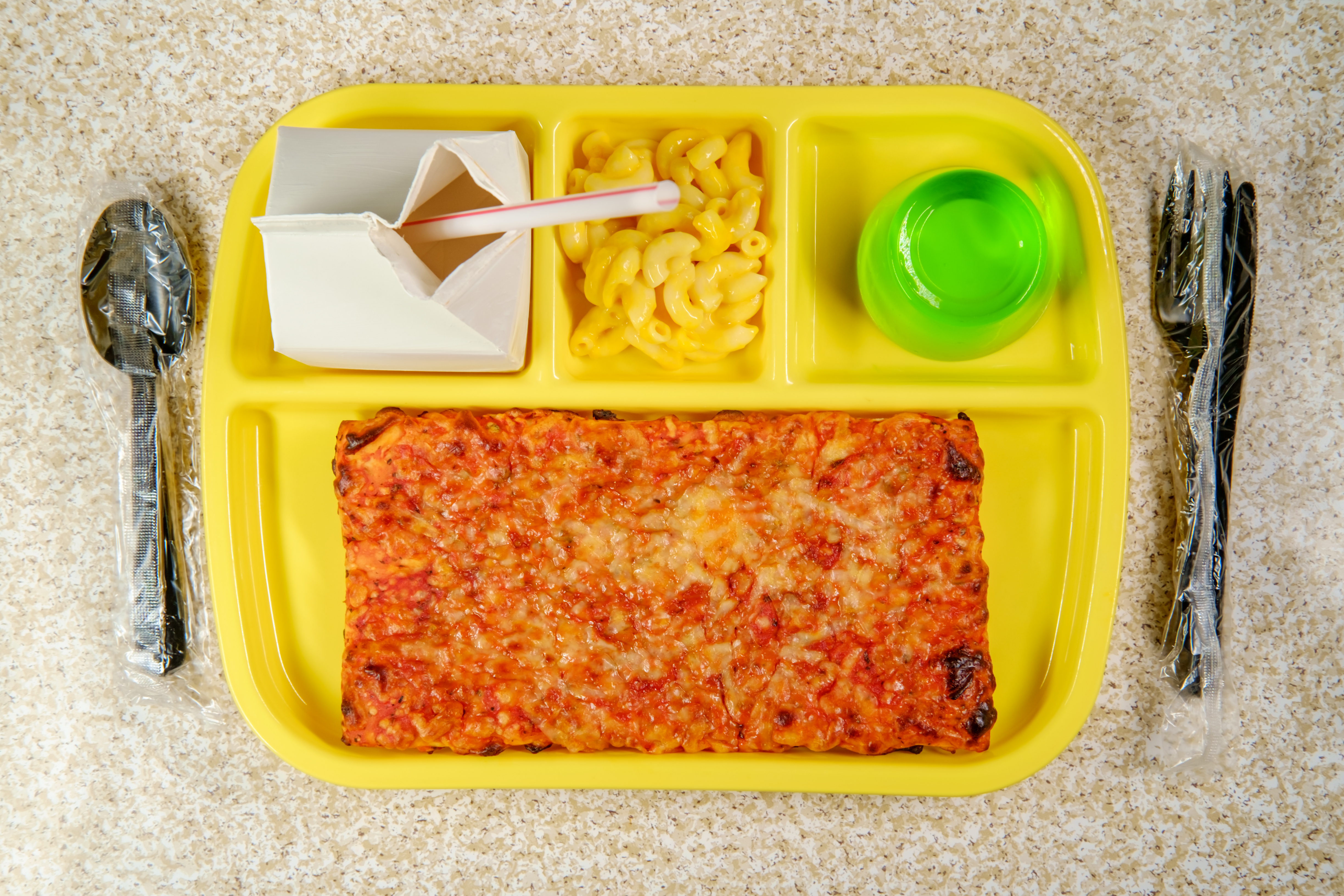 Bright yellow cafeteria tray with a square slab of pizza, open milk carton with straw, macaroni and cheese, and glass.