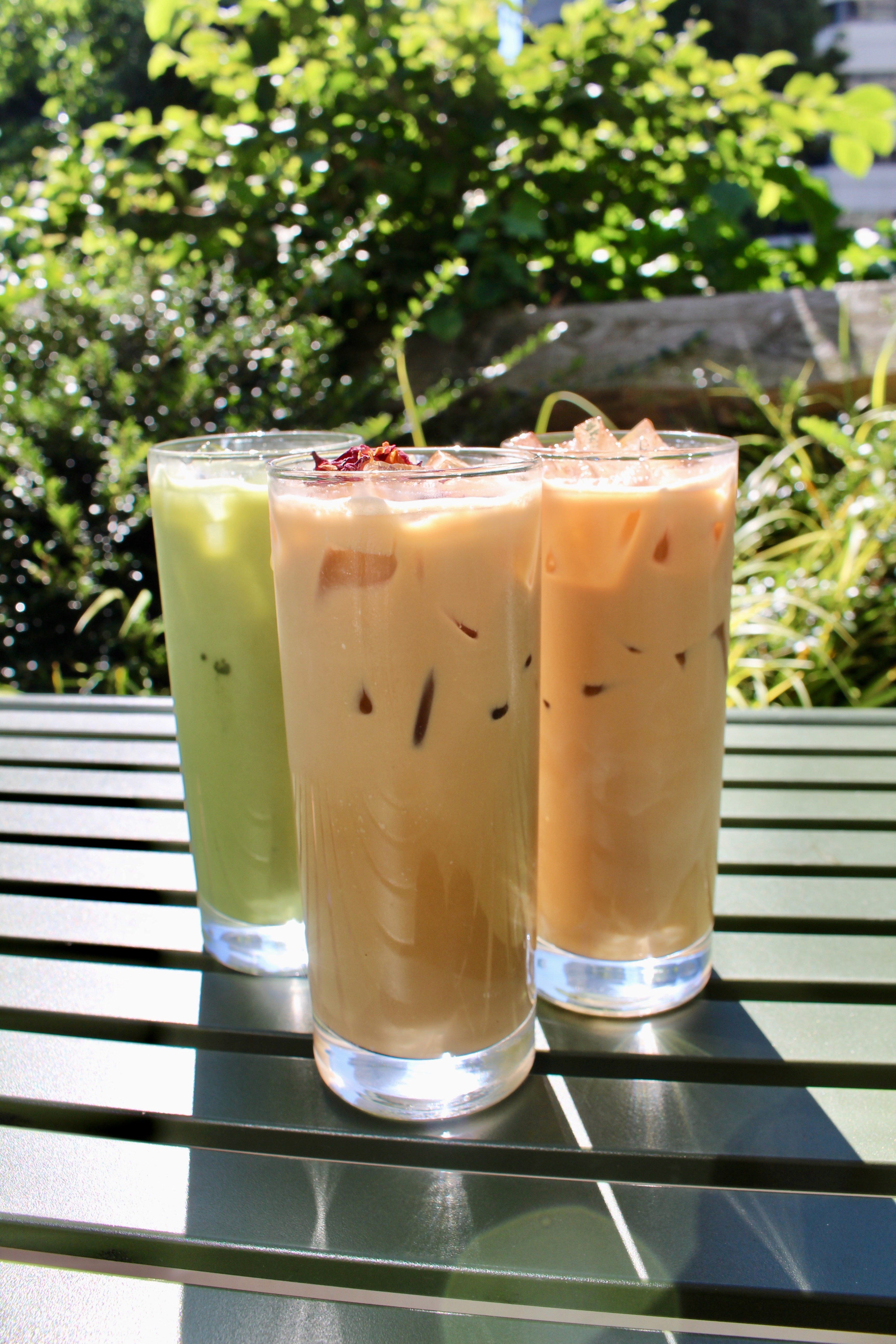Three tall glasses arranged in a triangle shape on a green metal outdoor table with horizontal slats. The sun shines brightly on the green shrubs growing in the background, light glinting off the ice cubes in the 3 lattes. From left to right: green iced lavender matcha latte, center is iced rose tahini latte glass that is coffee-colored with rose petal garnish floating on top, right is an iced oat milk lavender latte the color of coffee.