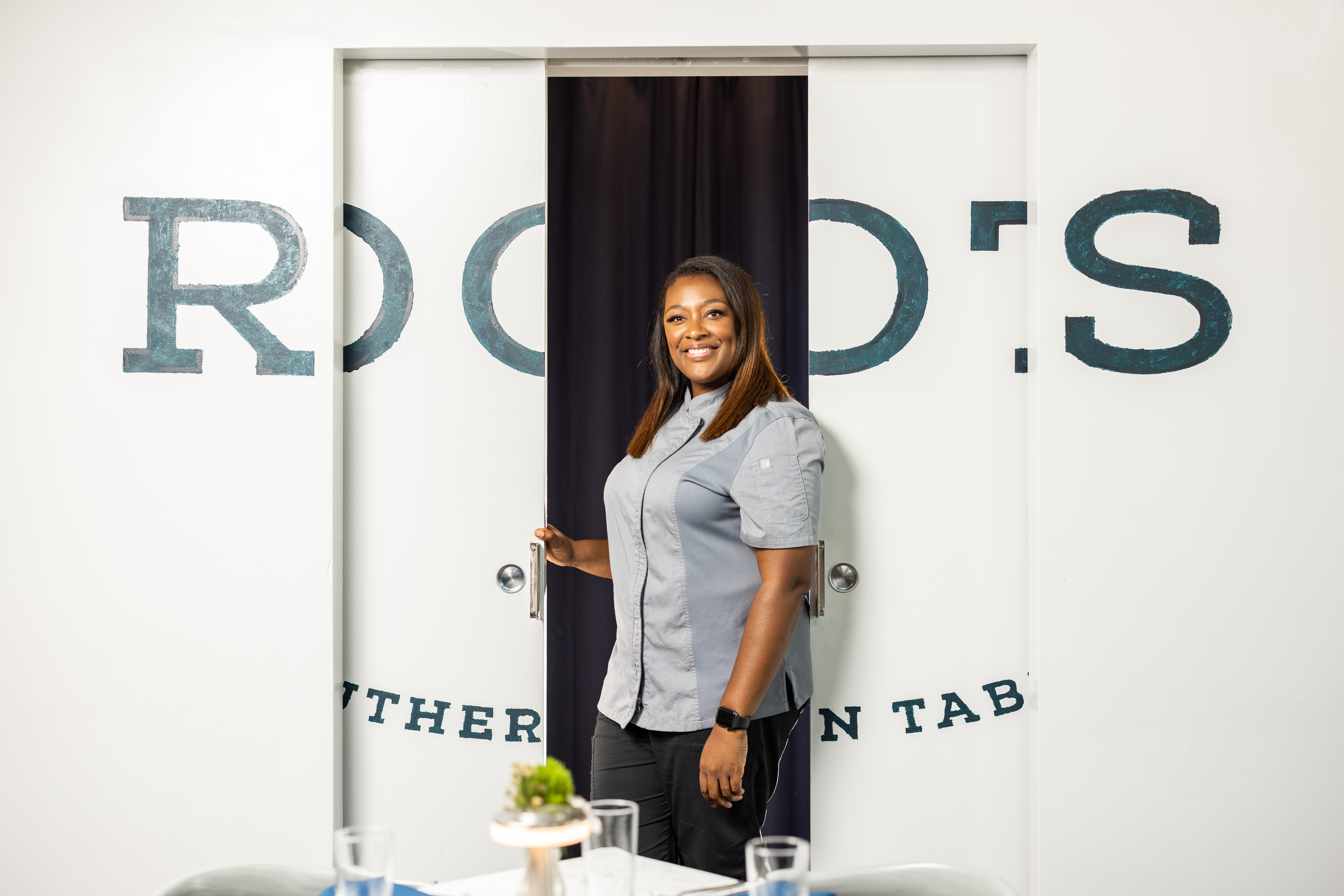 Chef Tiffany Derry stands in her chef gear in front of a sign that reeds “Roots Southern Table” in her Dallas restaurant.
