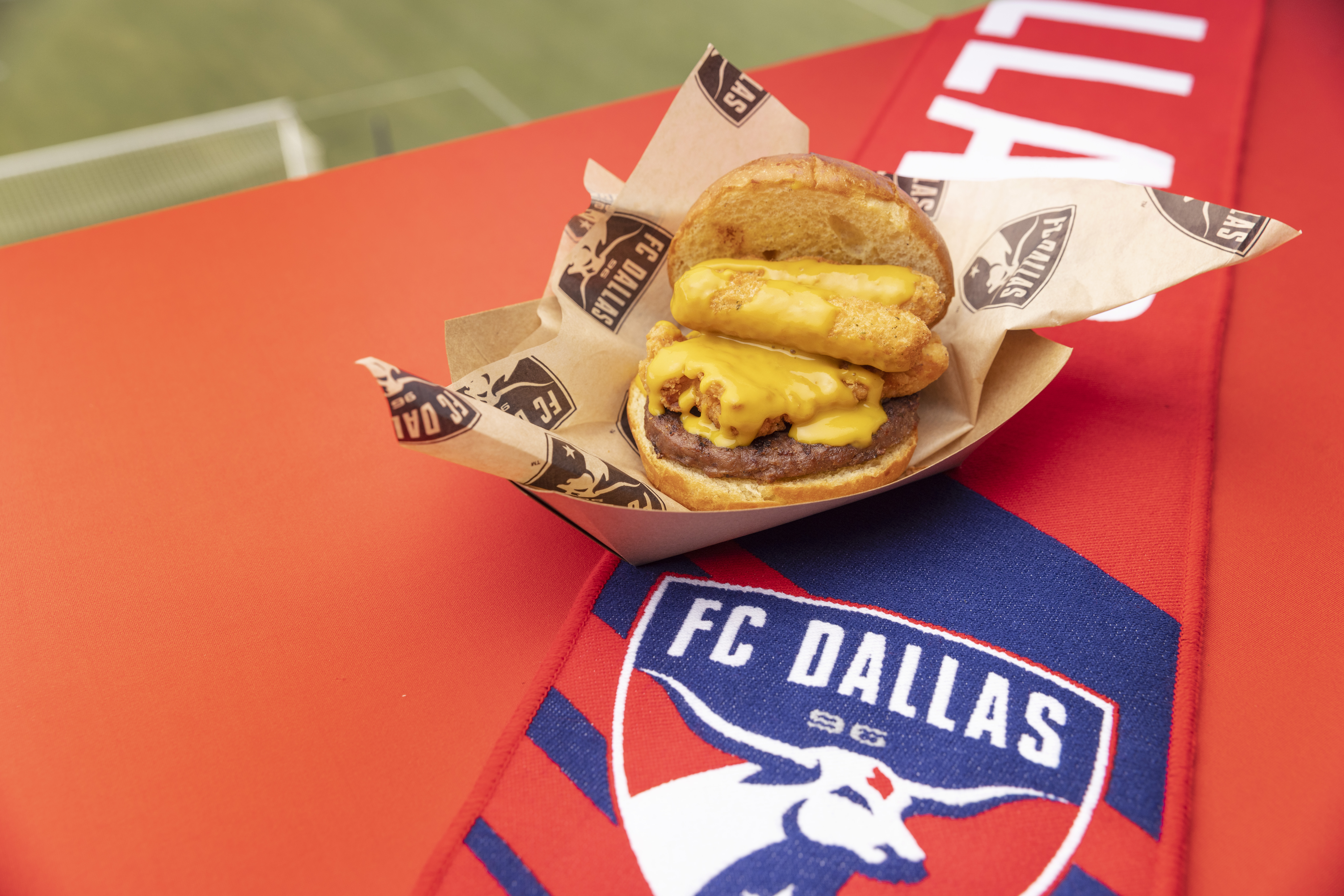 A burger topped with mozzarella sticks and honey mustard sits in a cardboard serving container, on top of a red background with the FC Dallas logo in blue and white.
