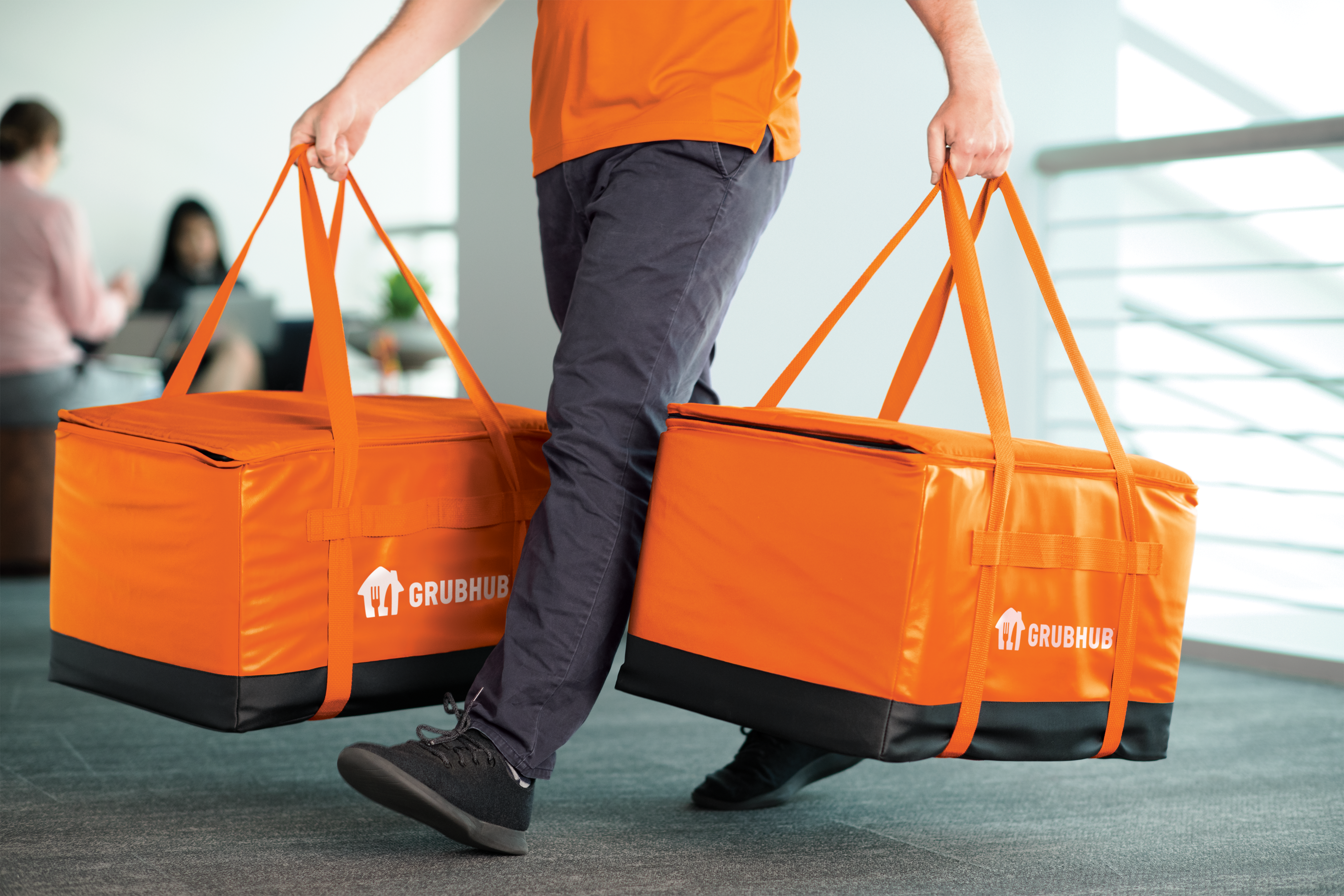 A person in dark jeans carrying two orange and insulated Grubhub delivery bags.