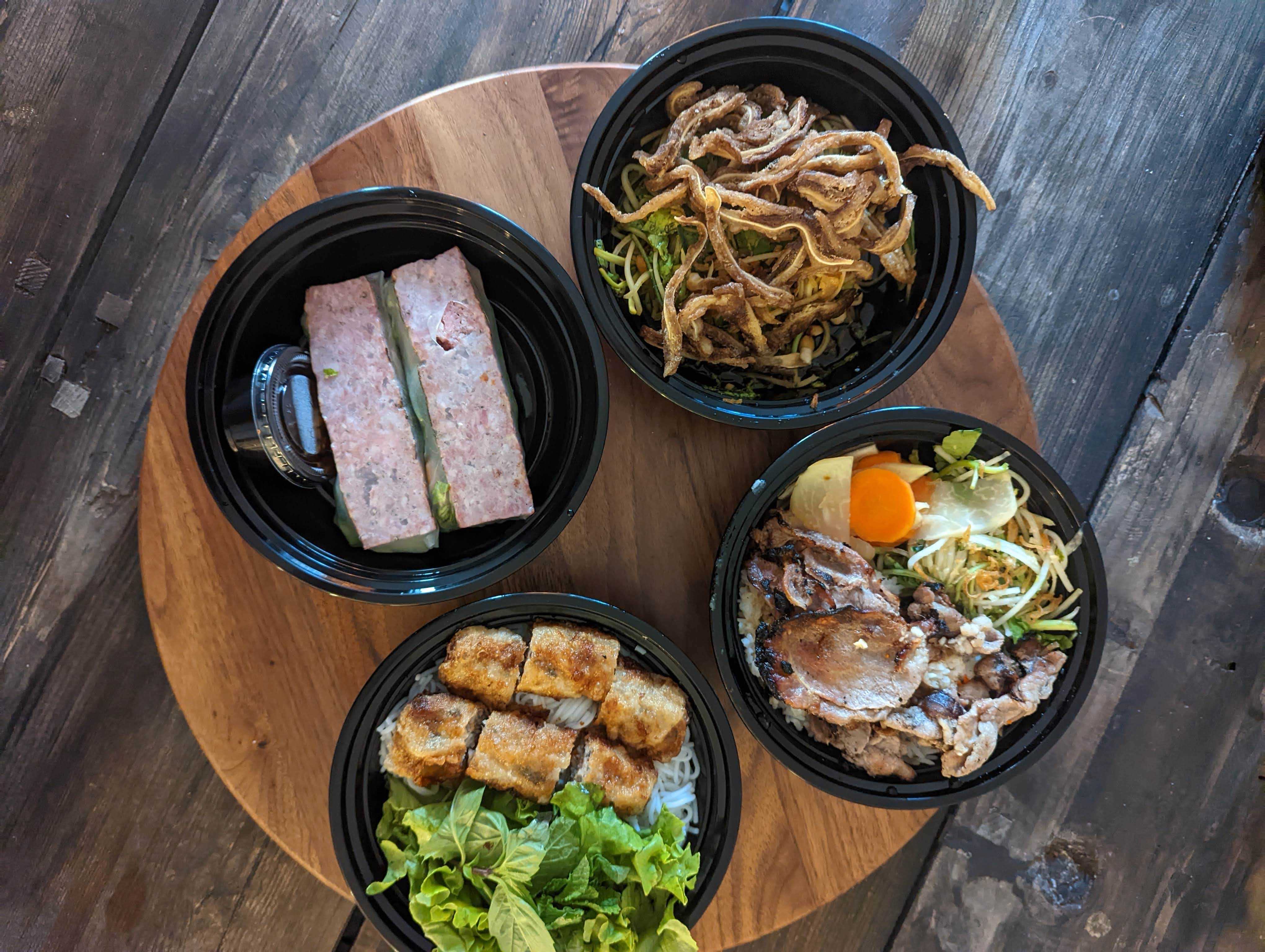 Four Vietnamese dishes in black plastic containers on a wooden table top.