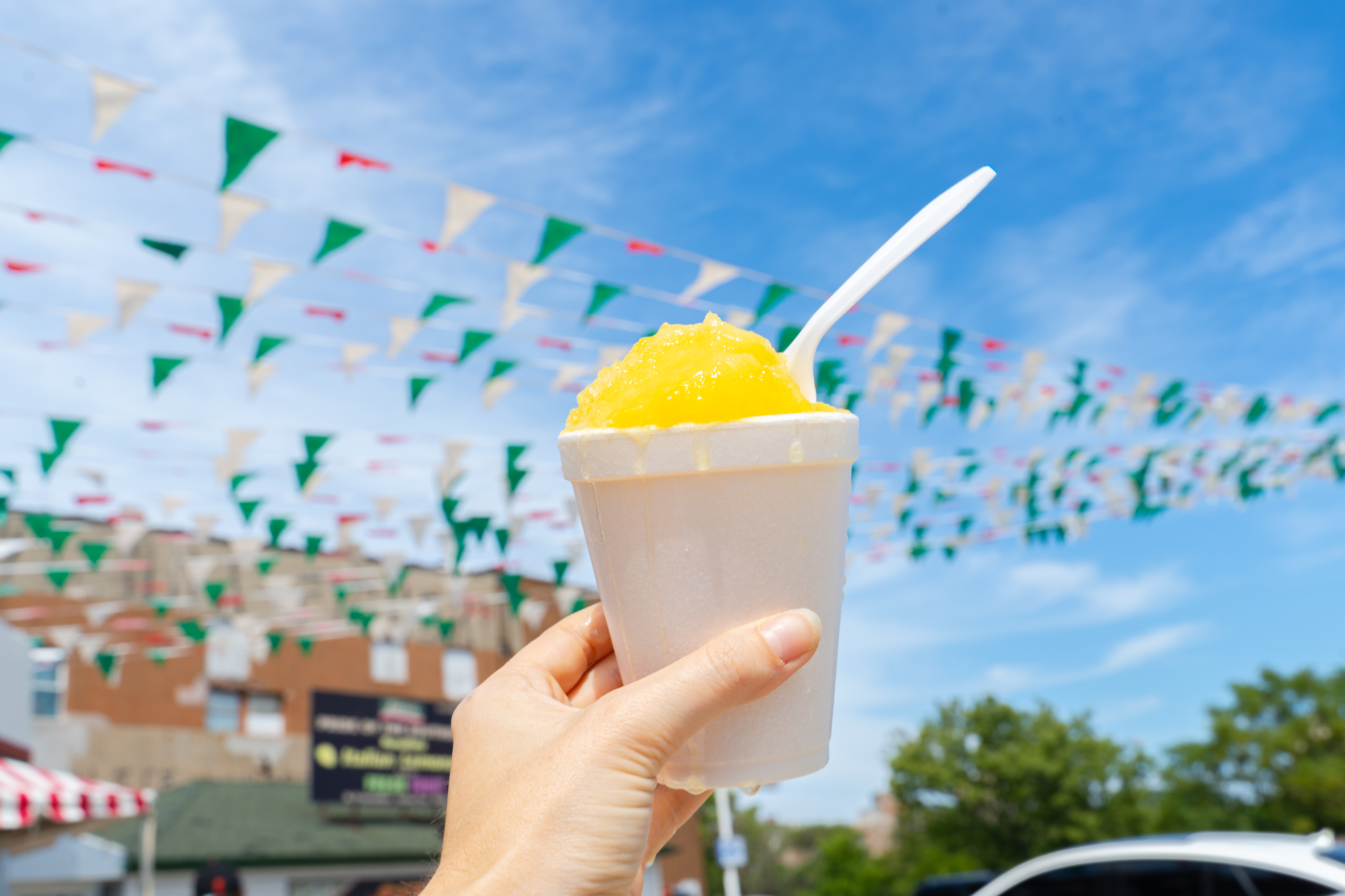 A hand holds up a white styrofoam cup filled with yellow Italian ice in front of a blue sky with white clouds and strings of red, green, and white flags.