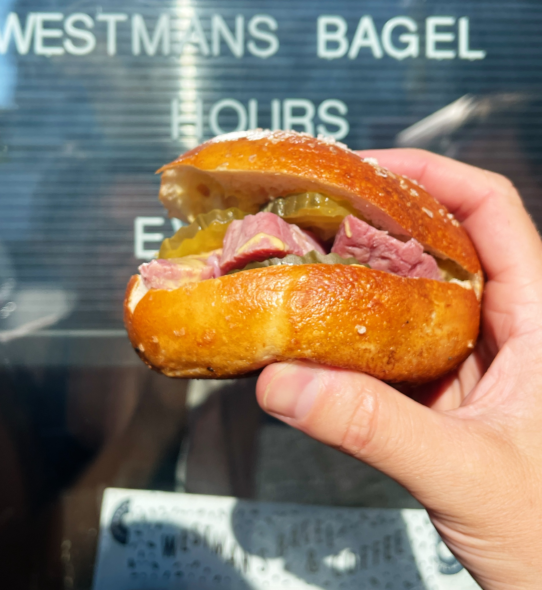 A bagel sandwich, held up by a hand, filled with pickles and corned beef.