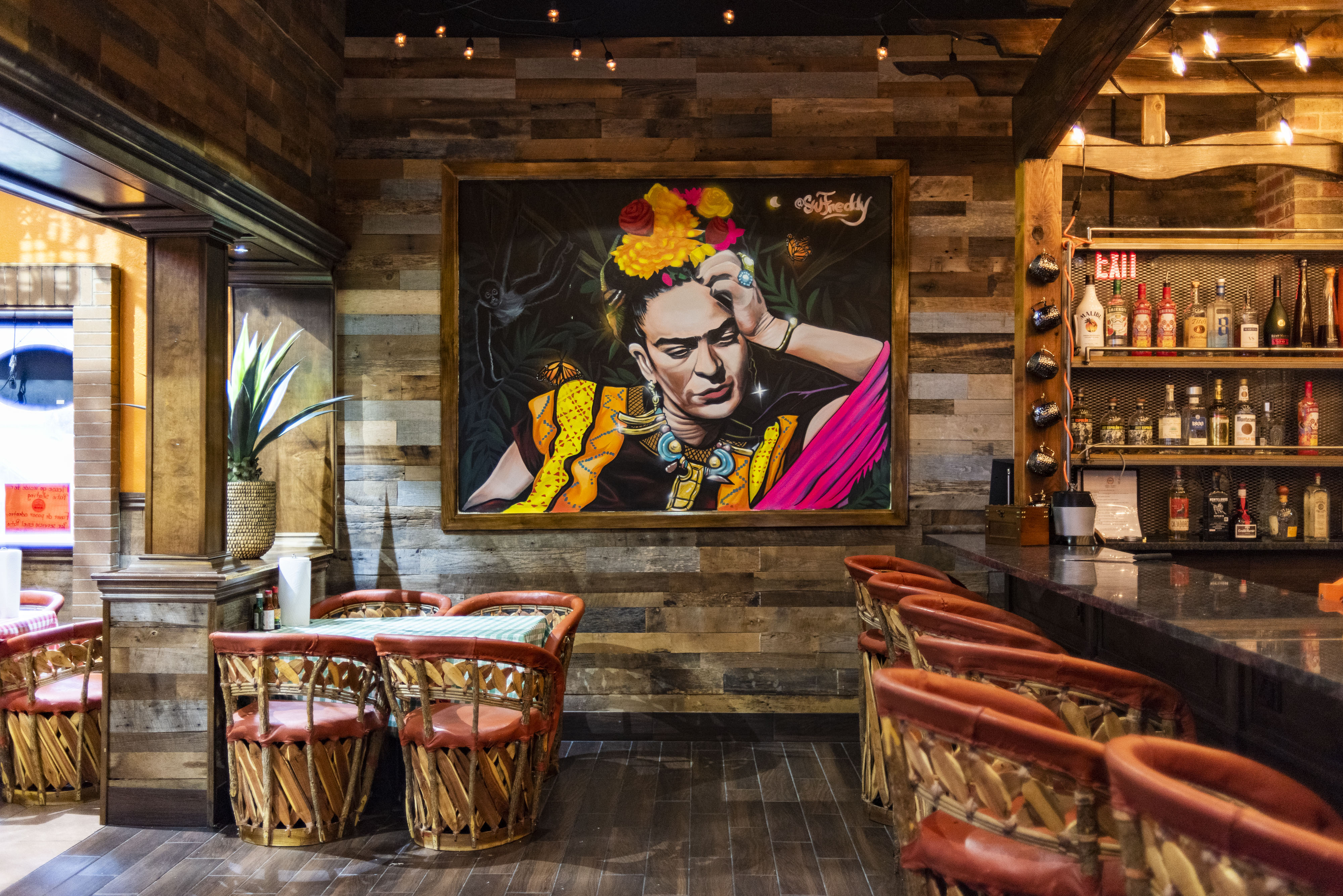 A painting of Frida Kahlo on a wood paneled wall with several chairs and a bar.