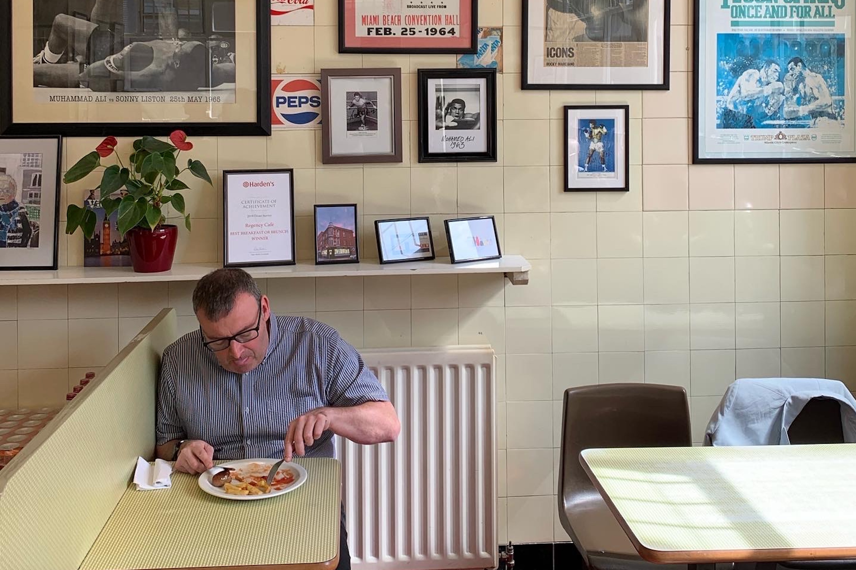 A man sits at a table in a London caff, eating a fry-up off a white plate. Behind him are posters and framed clippings from history.