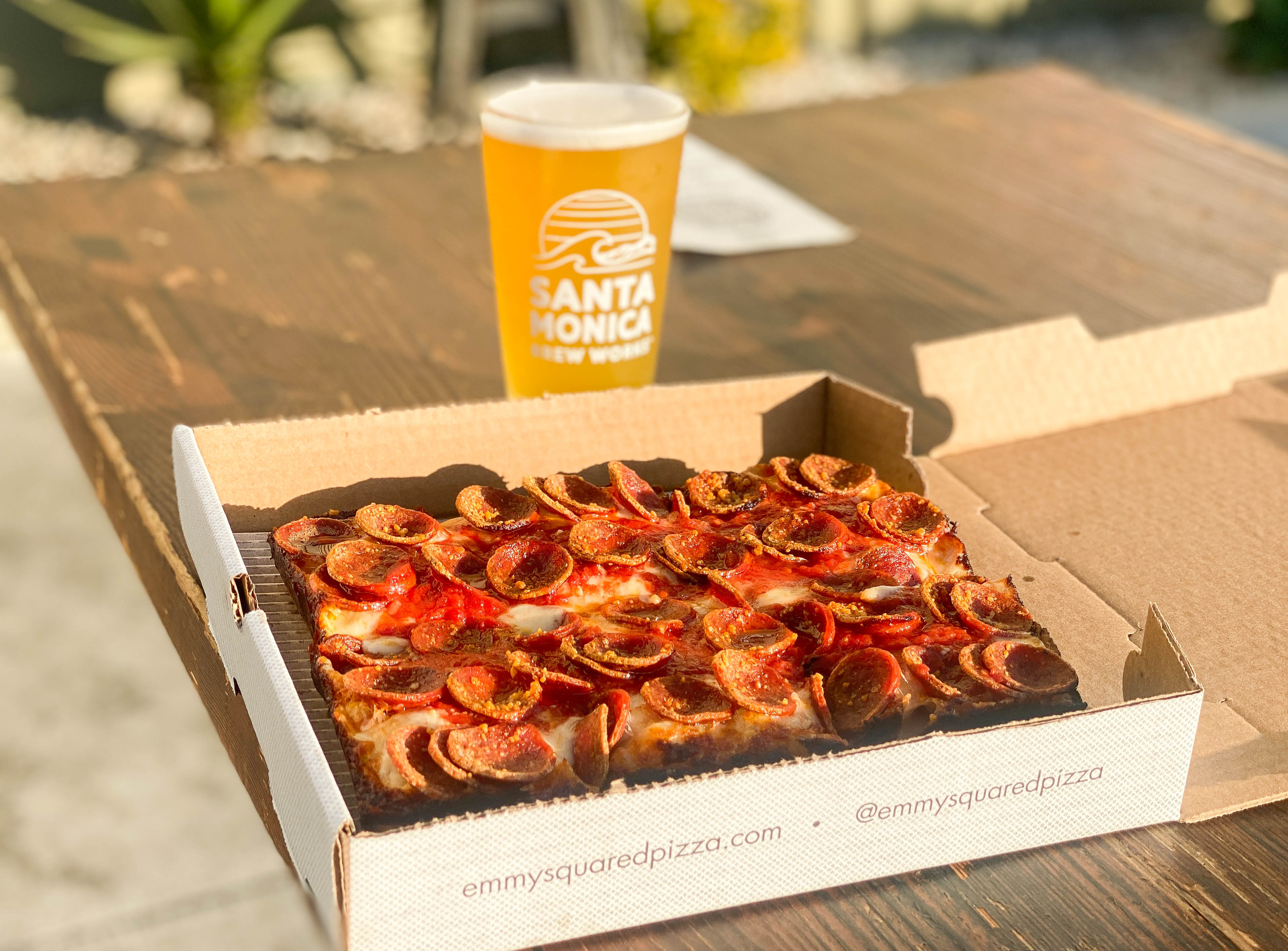 A square pizza in a to go box with a pint of light yellow beer beyond.