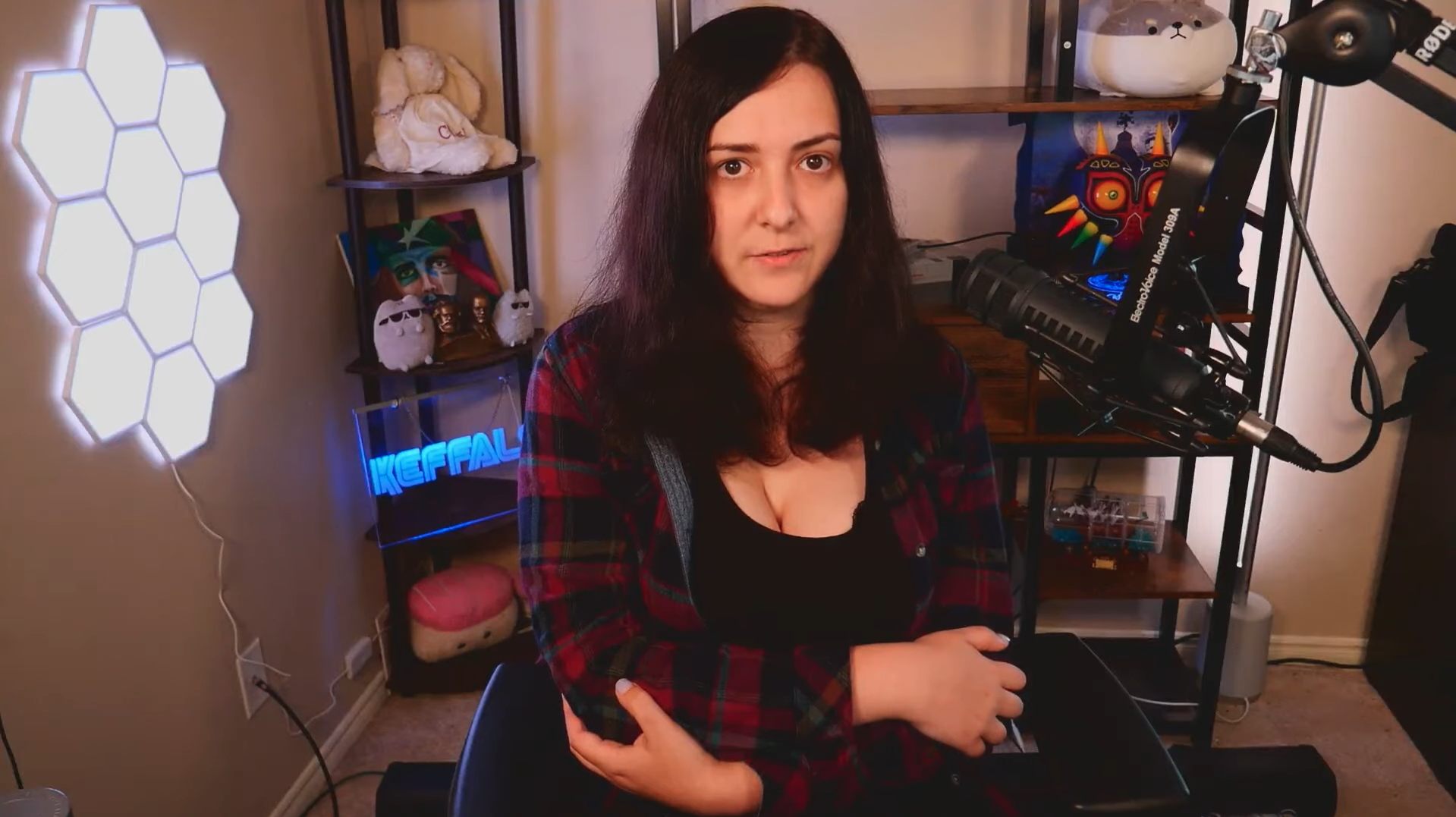 a still from a video from the streamer Keffals sitting at a gaming setup. Their posture is reserved and they’re holding their arms hugged in as they look into the camera. 