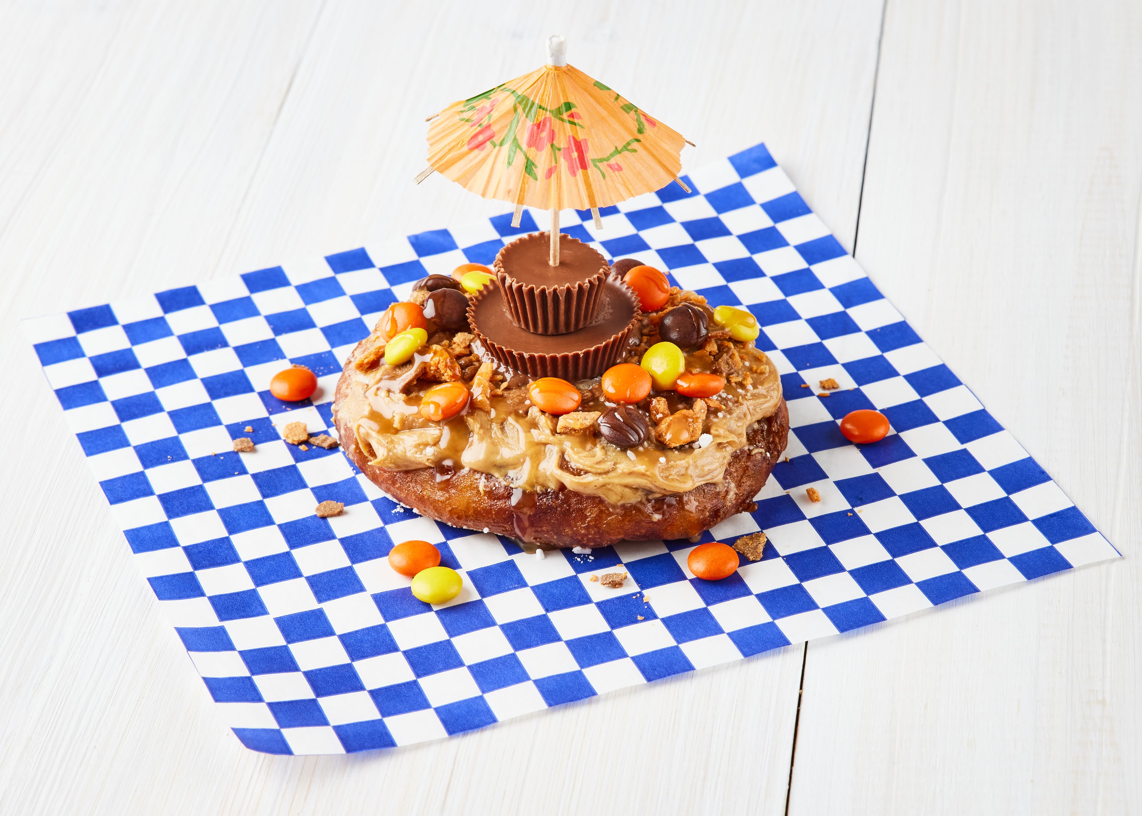 A cake sits on a blue and white checked napkin. It’s covered with peanut butter, Reeses Pieces, and two Reeses Peanut Butter Cups, with a cocktail umbrella on top.