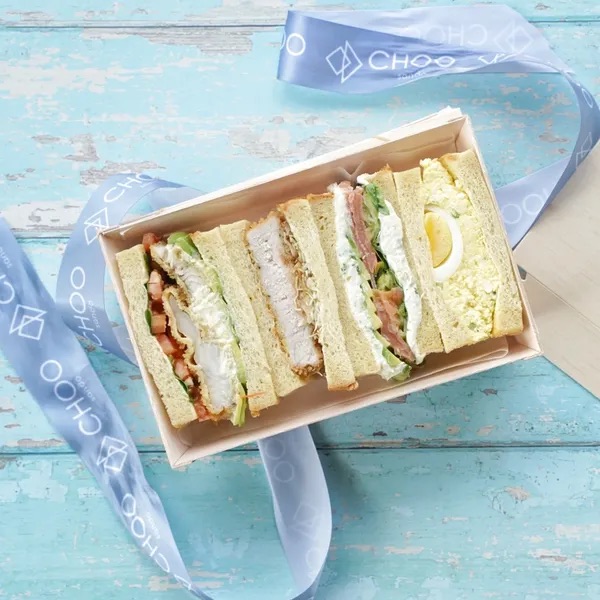 A box of four kinds of Japanese sandwiches on a blue table with ribbon next to it that says Choo Sando