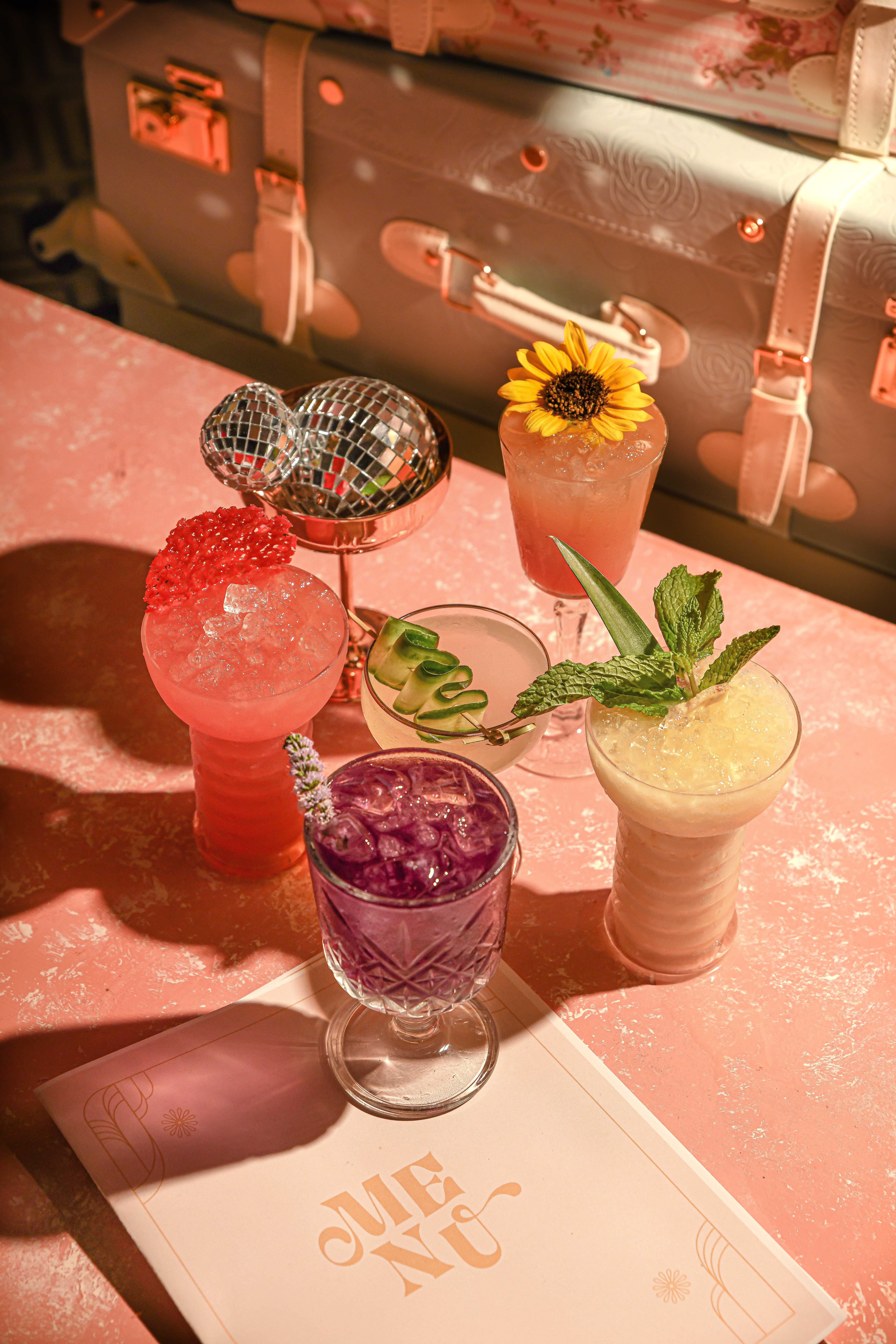 Five colorful cocktails sit on a table covered with a pink and white top. In the upper left corner, a tabletop sized disco ball sits with them.