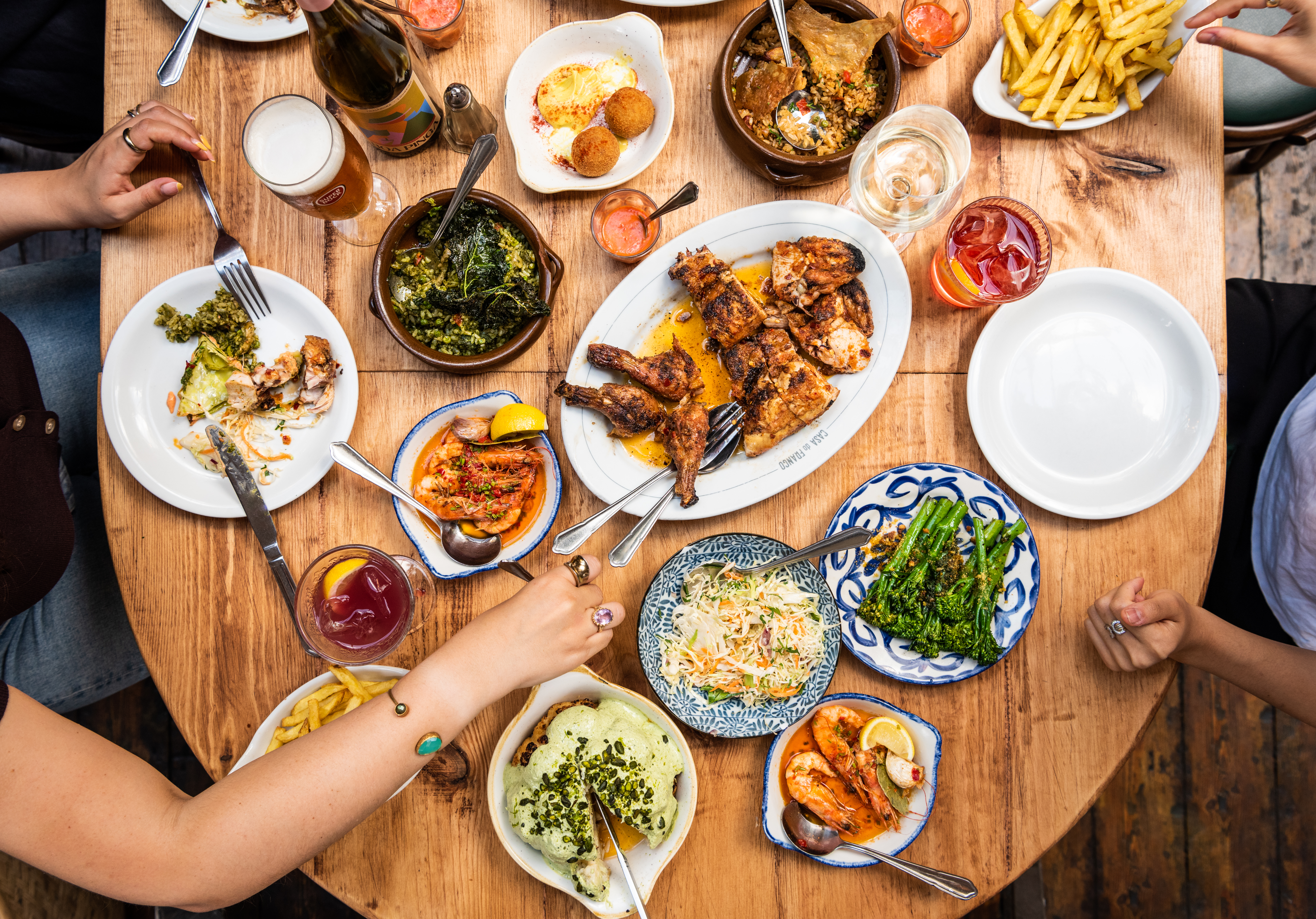 A birdseye shot of a wooden table full of food, including platters of piri piri chicken, grilled broccoli, croquettes, and chips, with people digging in.