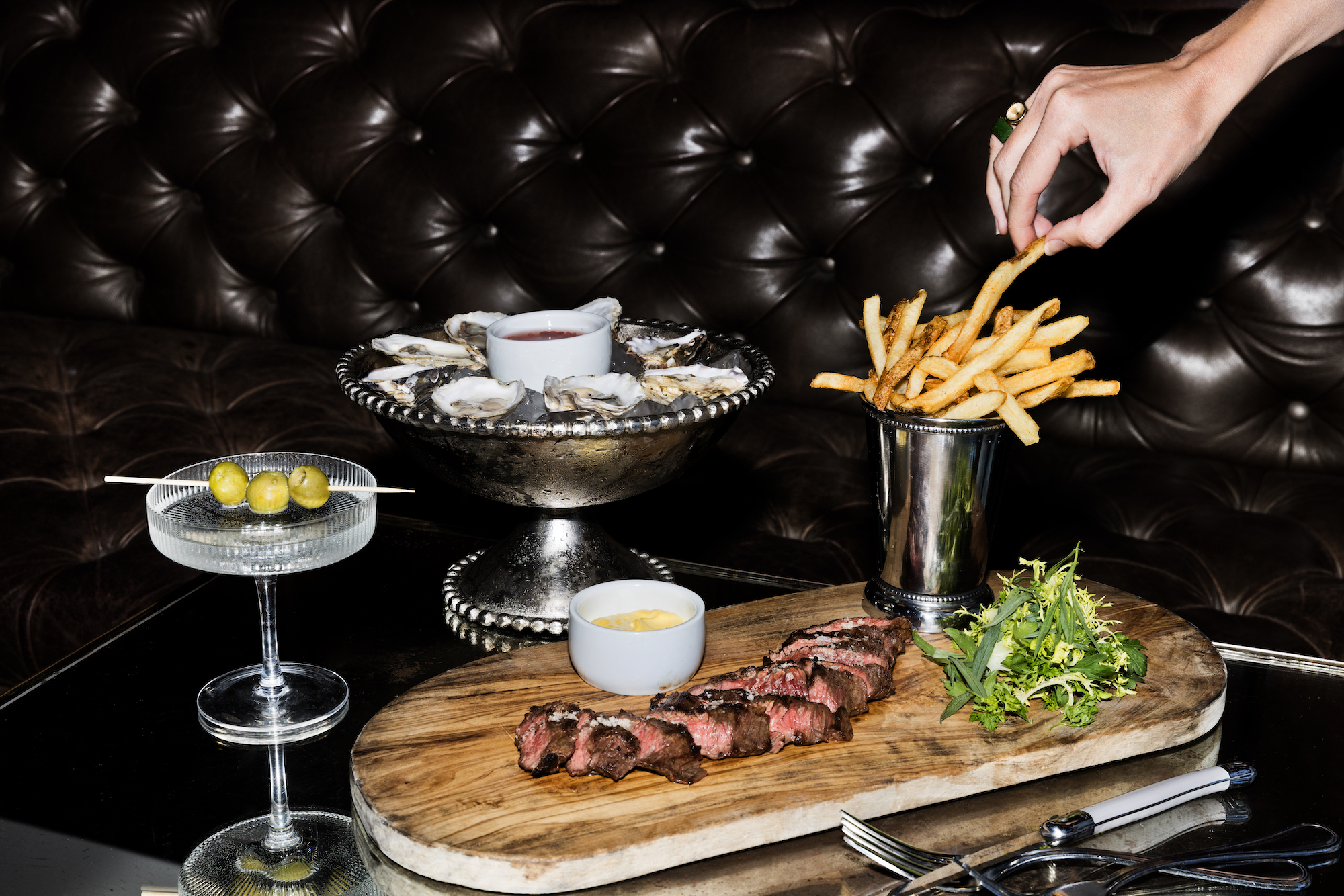 A hand grabs french fries as a flash goes off next to a steak inside a dark restaurant with black leather booths.