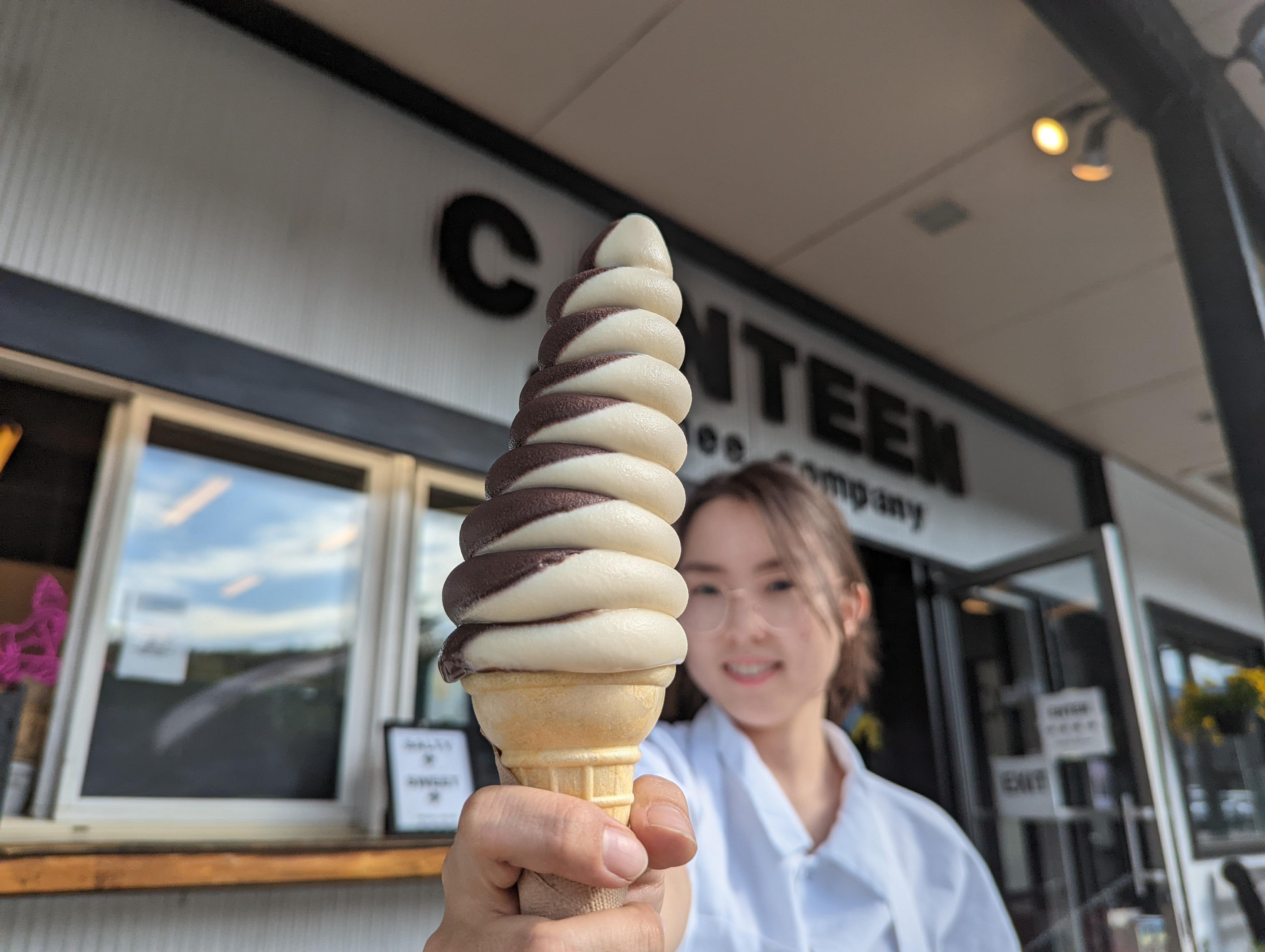 A staff member holds out a tall swirl creemee in a cone, standing in front of a blurred storefront. 