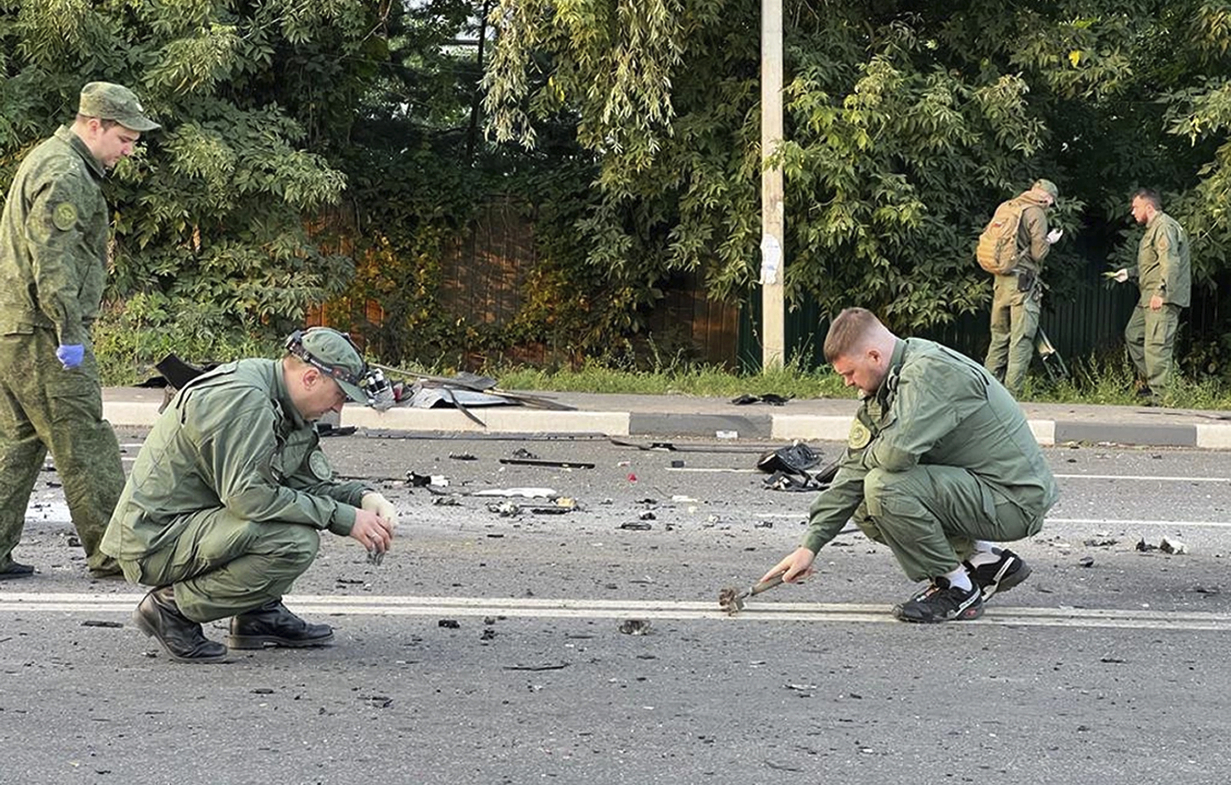 People in green jumpsuits crouch on a roadway looking at debris.