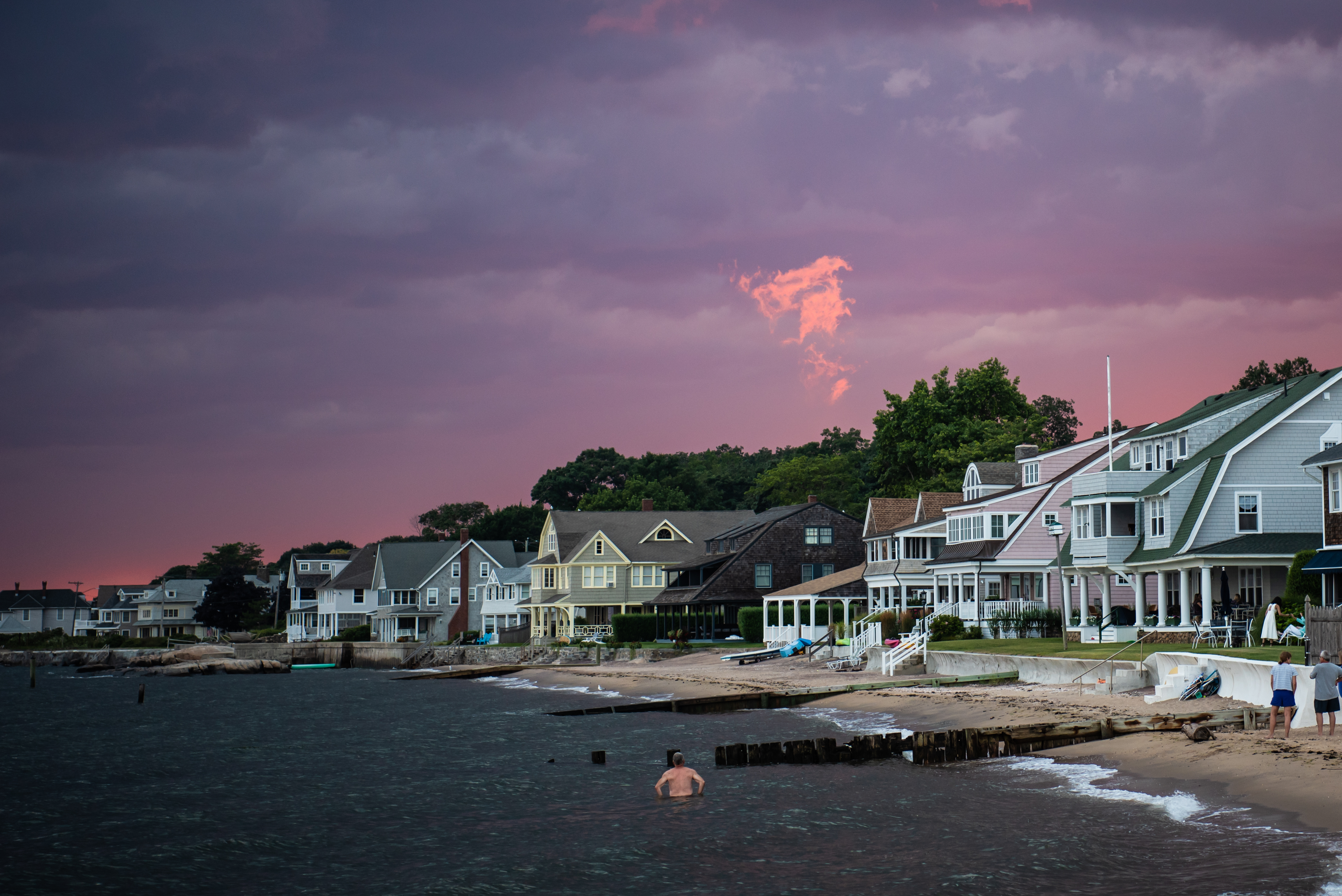 The sky is dark blue with a bit of pink above a shoreline, with houses lining the beach just feet from the water.