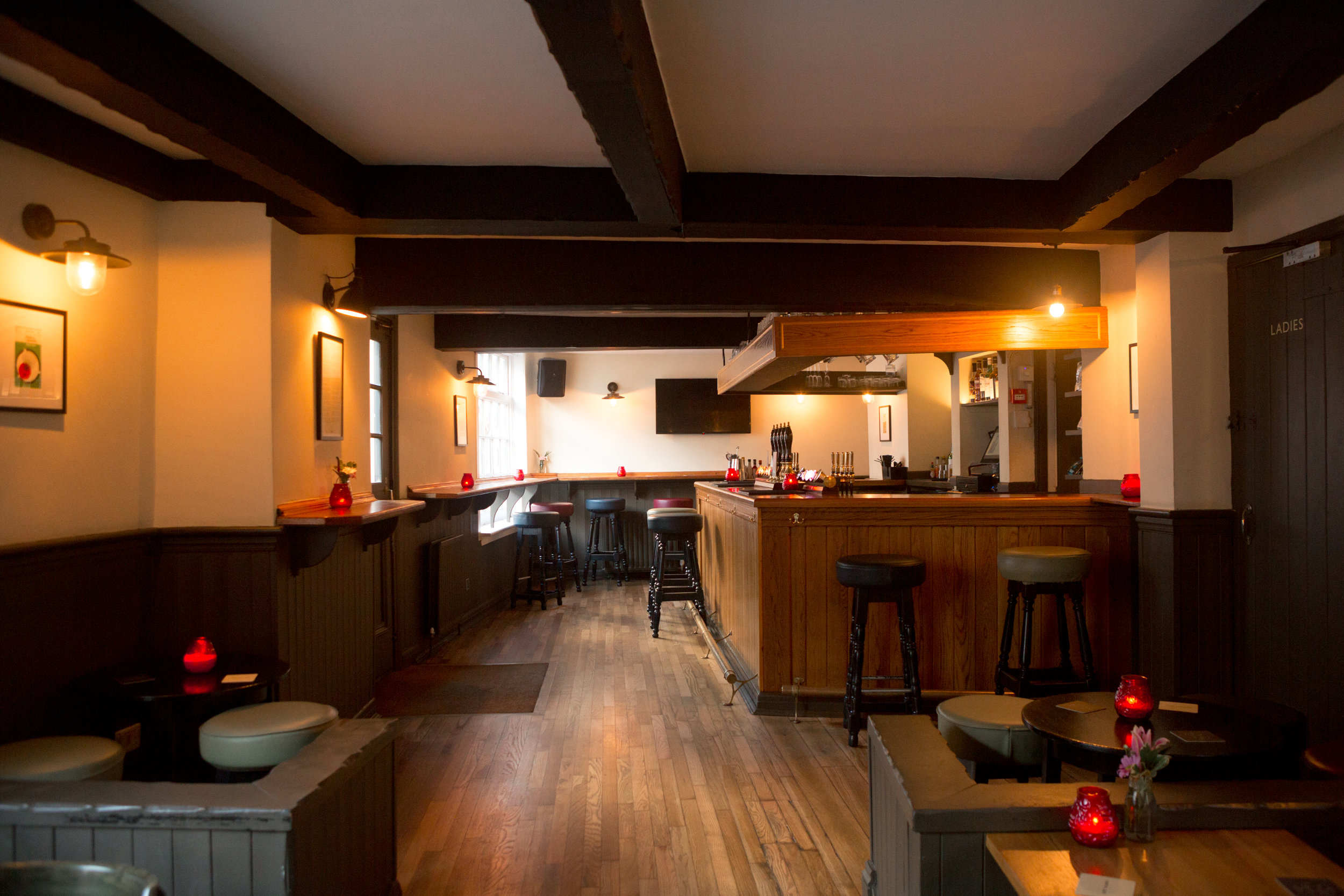 A wood-panelled pub interior, with the bar to the right in the background and tables and stools in front.