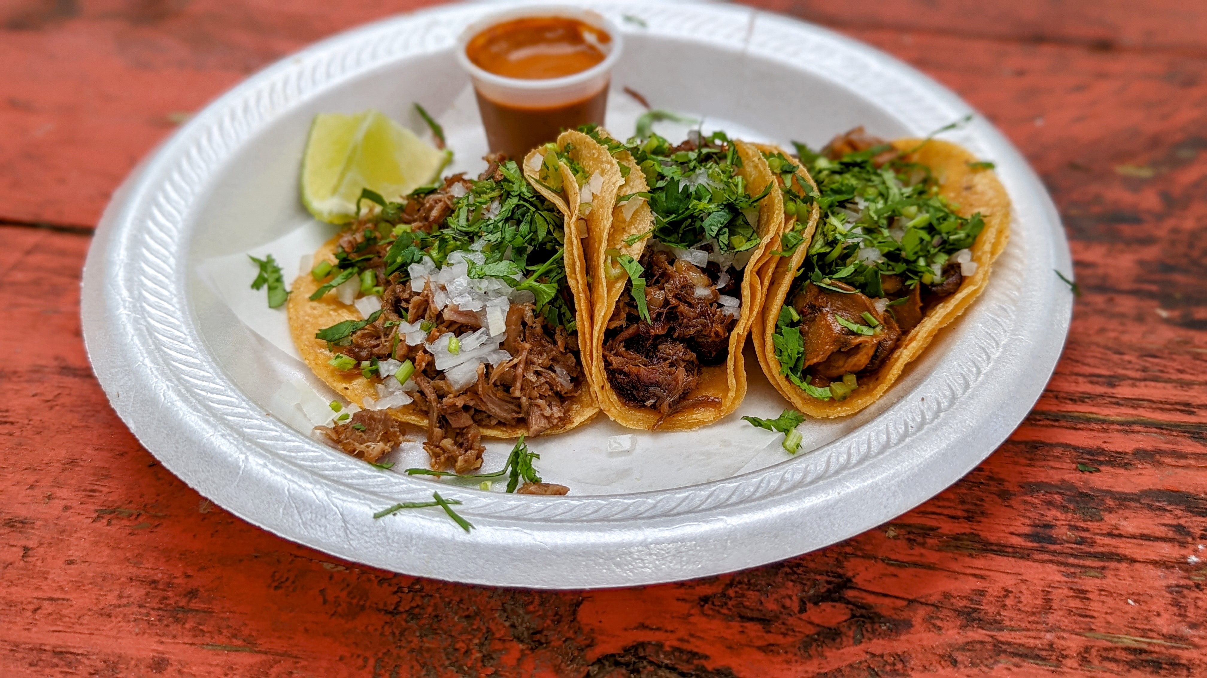Three tacos full of meats and fillings on a white styrofoam plate on a reddish wood table. 