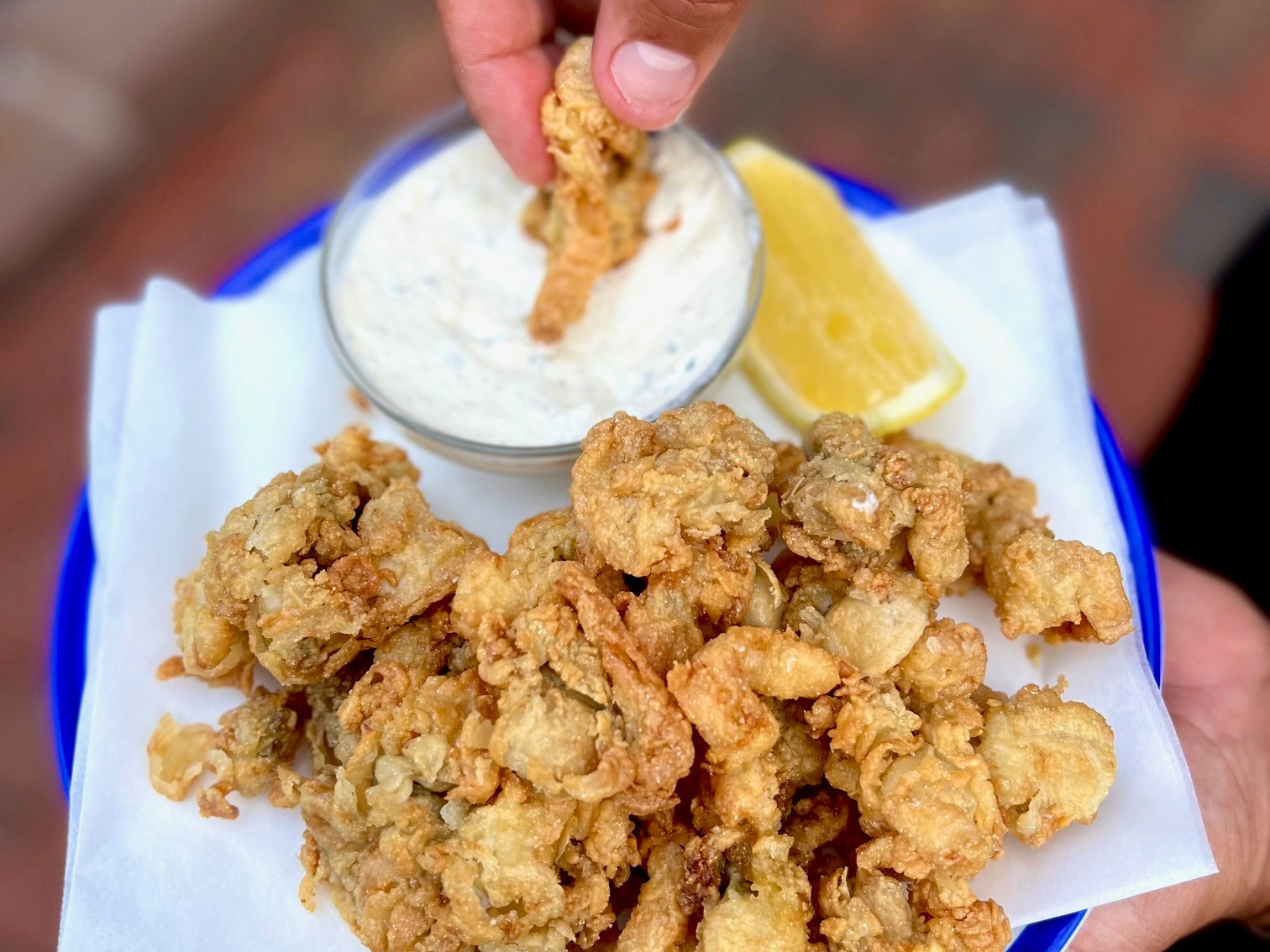 A hand dips a fried clam into a cup of tartar sauce, surrounded by more pieces of fried clam and a lemon wedge.