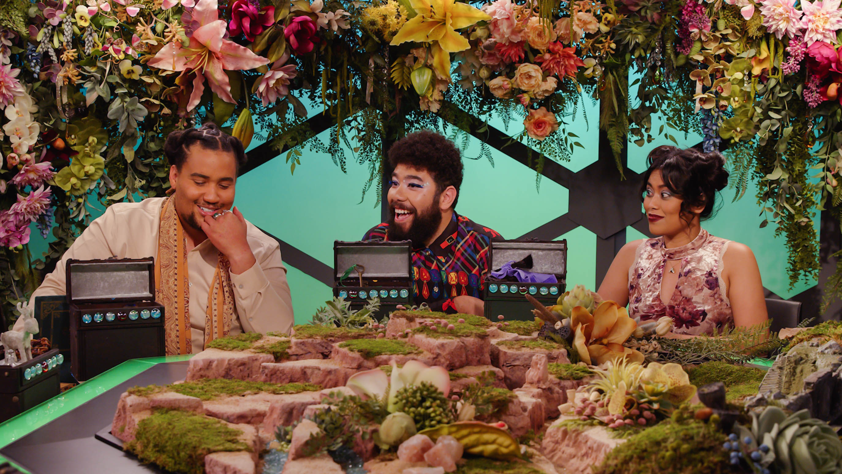 Three players at an elaborate board game table, covered in moss, flowers, and rock formations. Garlands hang above their heads.