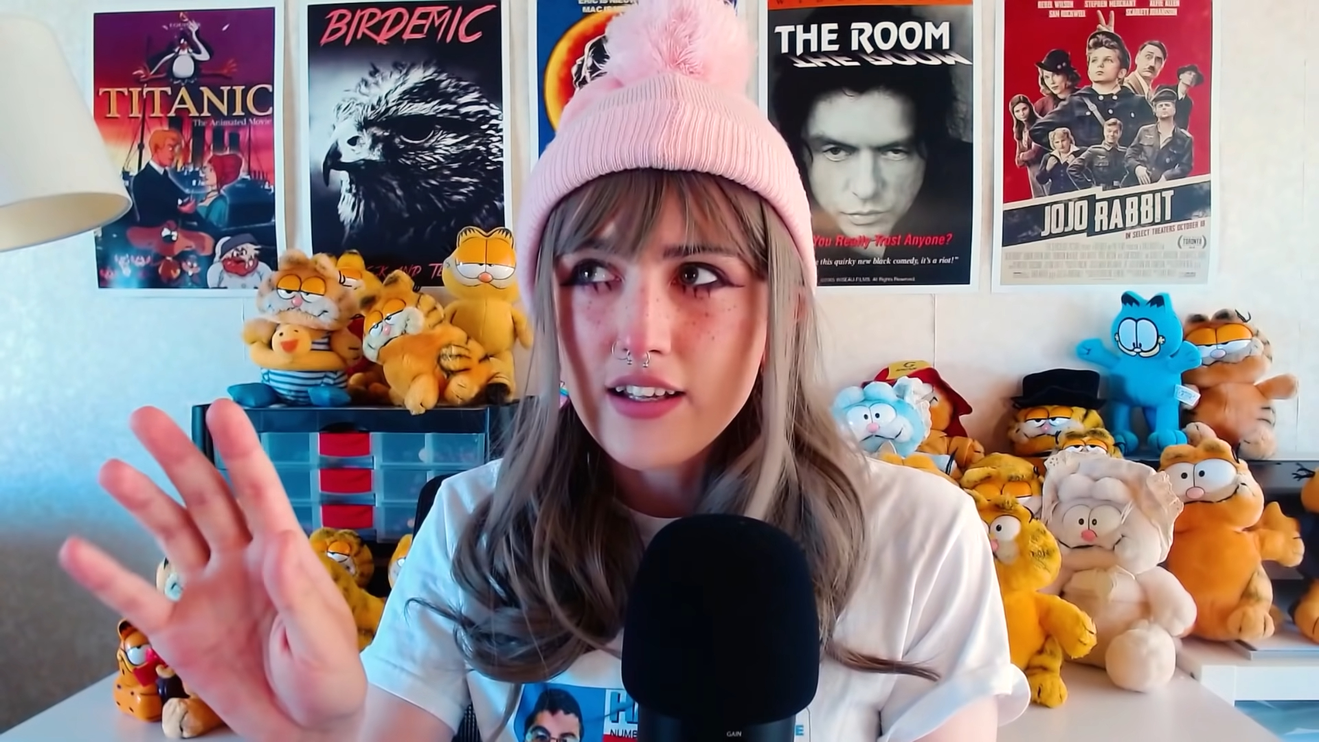 A young person wearing cute eye makeup and a pink beanie, looking to the left and holding up a hand, in a room with lots of Garfield plushies and some movie posters in the background.