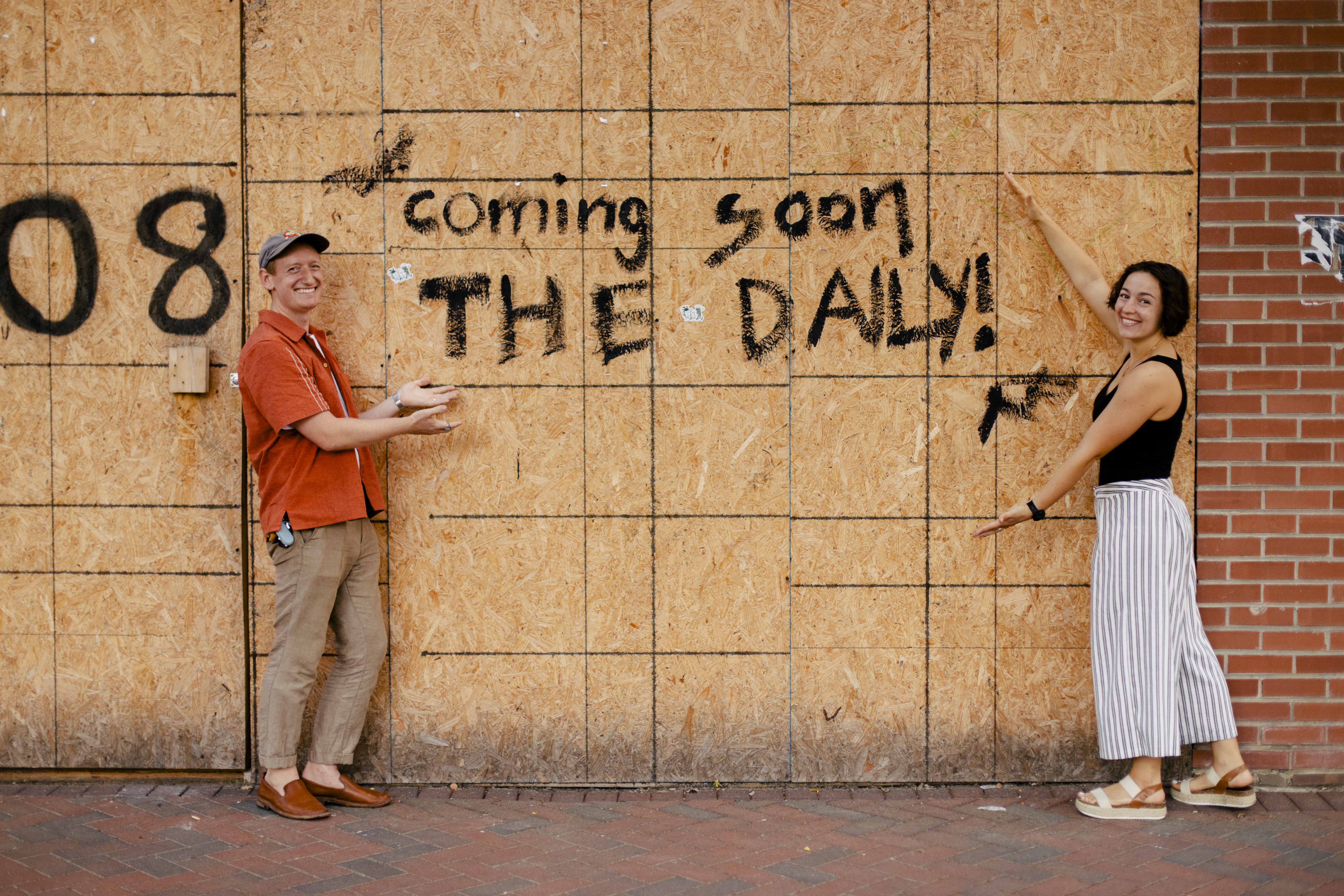 A man and a woman in front of plywood sign saying “coming soon.” 
