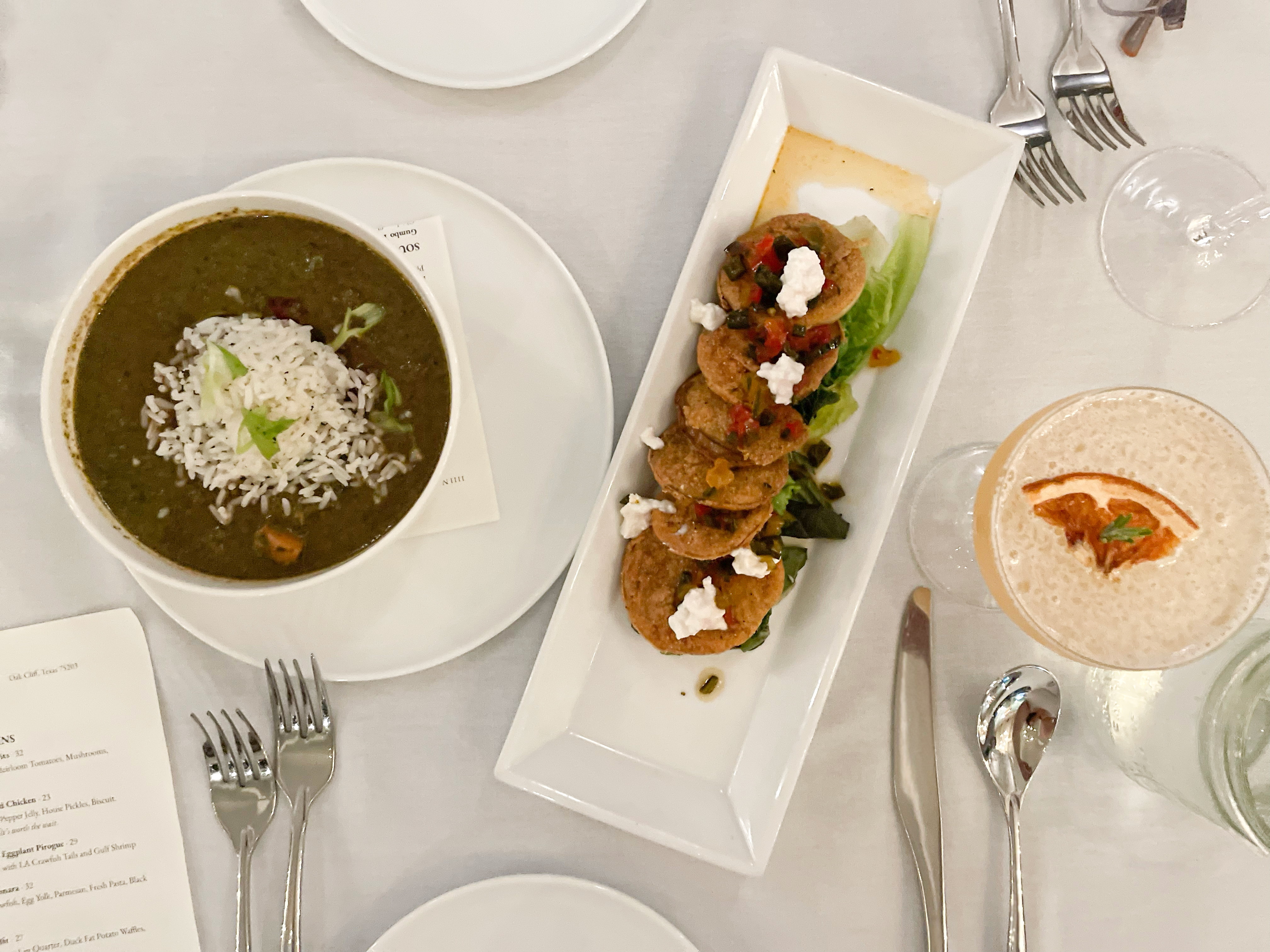 On table cover with a white tablecloth, set with silverware, a bow of gumbo sits to the left. In the middle is a tray dish of fried green tomatoes. And on the right is a light orange cocktail with a dried orange floating in it.