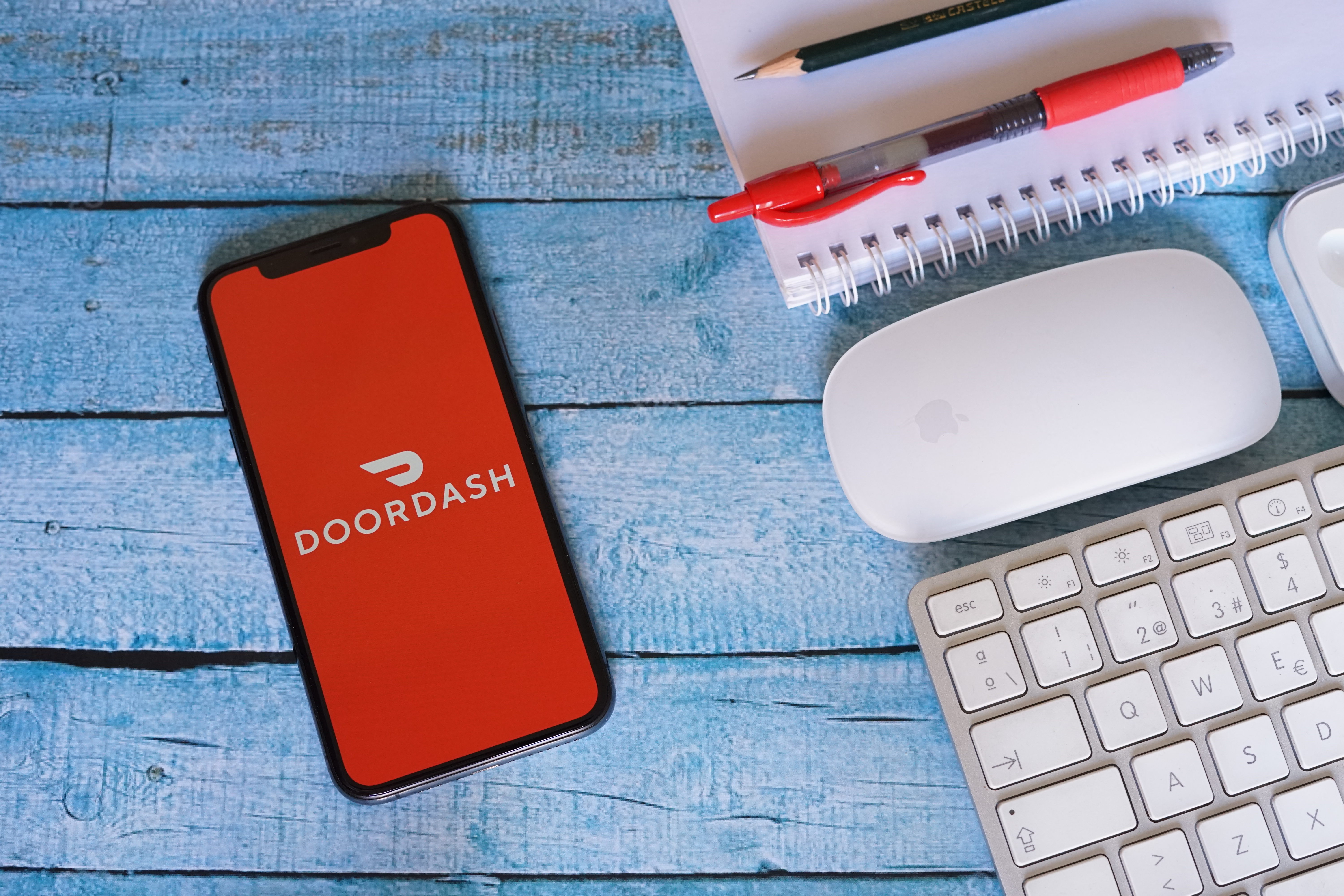 A cell phone showing the DoorDash app sits on a table next to a mouse, keyboard, and notebook.