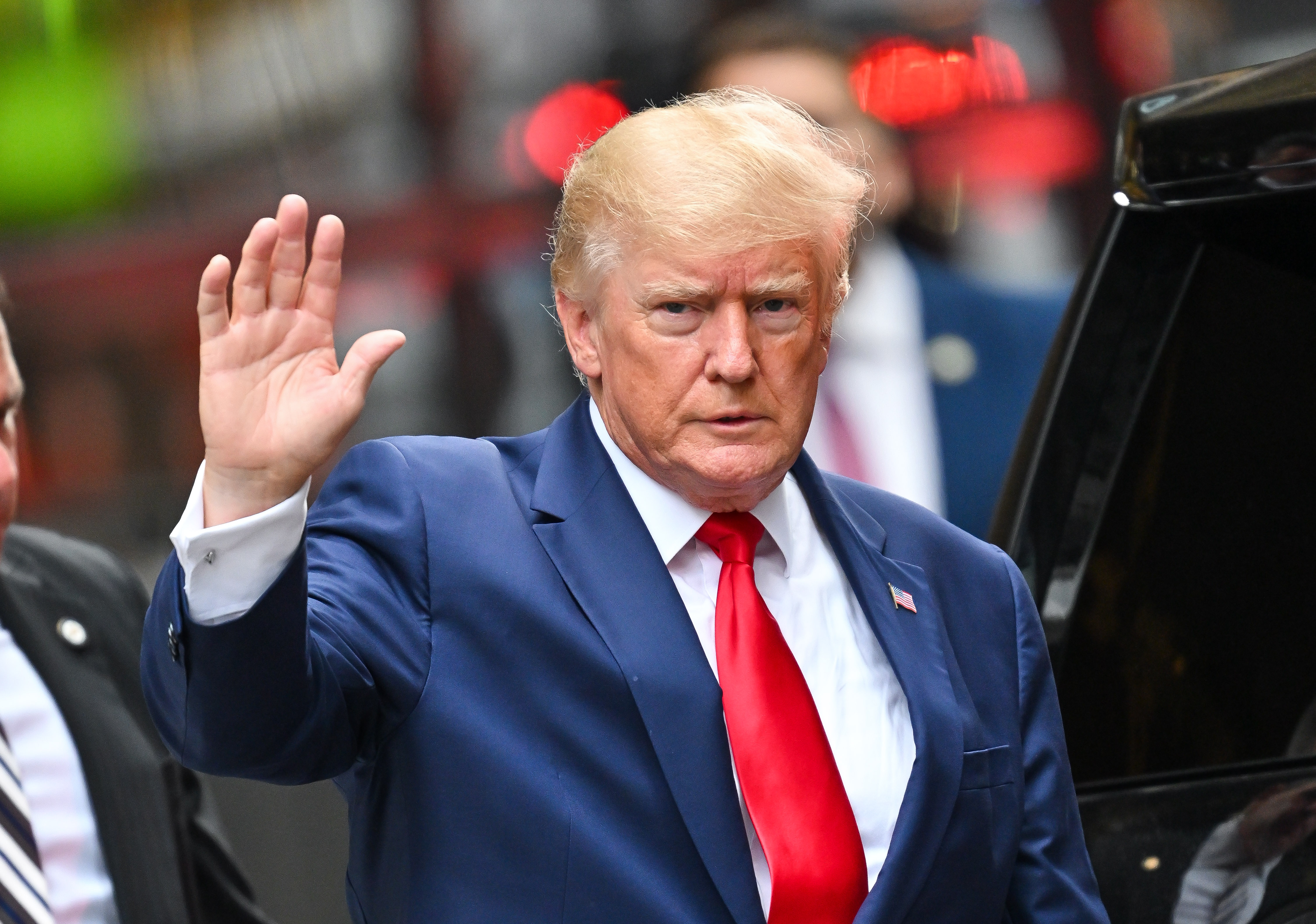 Donald Trump is pictured in a navy business suit with red tie. He is waving his right hand while looking into the camera.